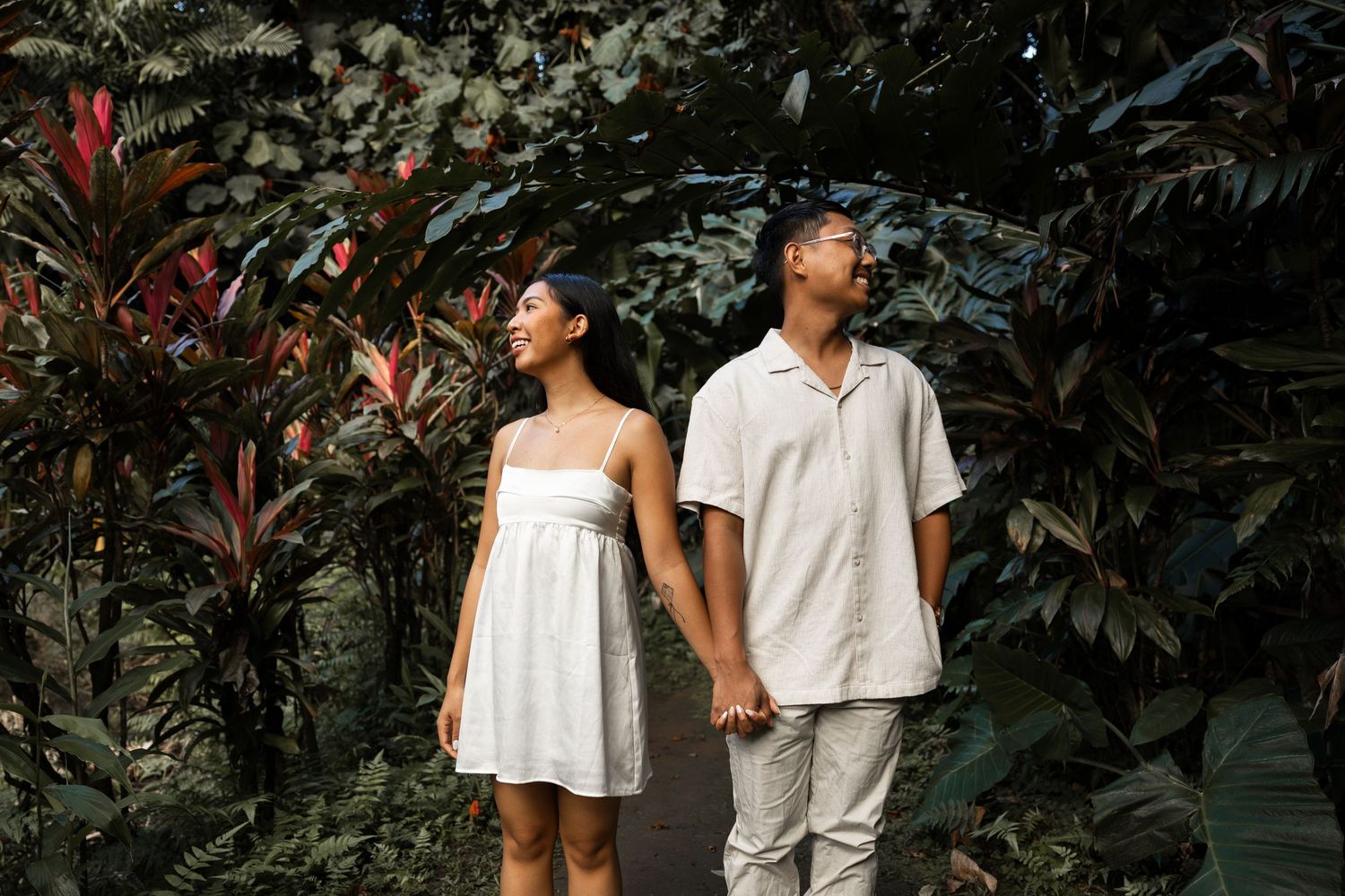 A couple in white outfits poses together in a lush tropical garden setting with dark green foliage.