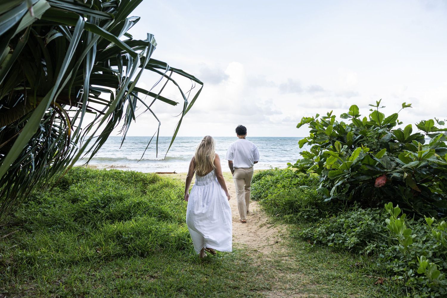 A couple walks down a tropical path towards the ocean with lush vegetation surrounding them.