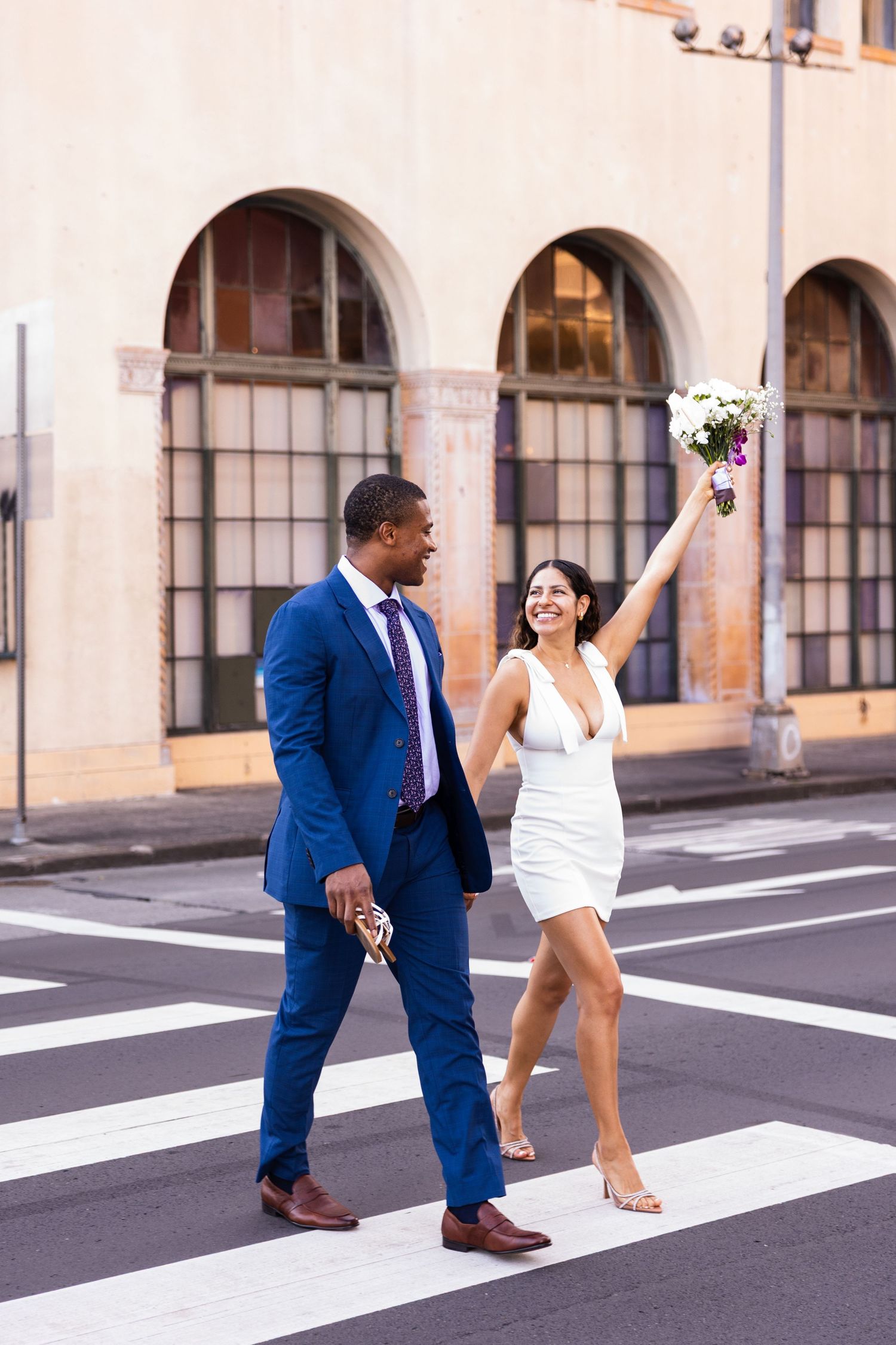A joyful couple walks across a street as one holds up a bouquet in celebration.