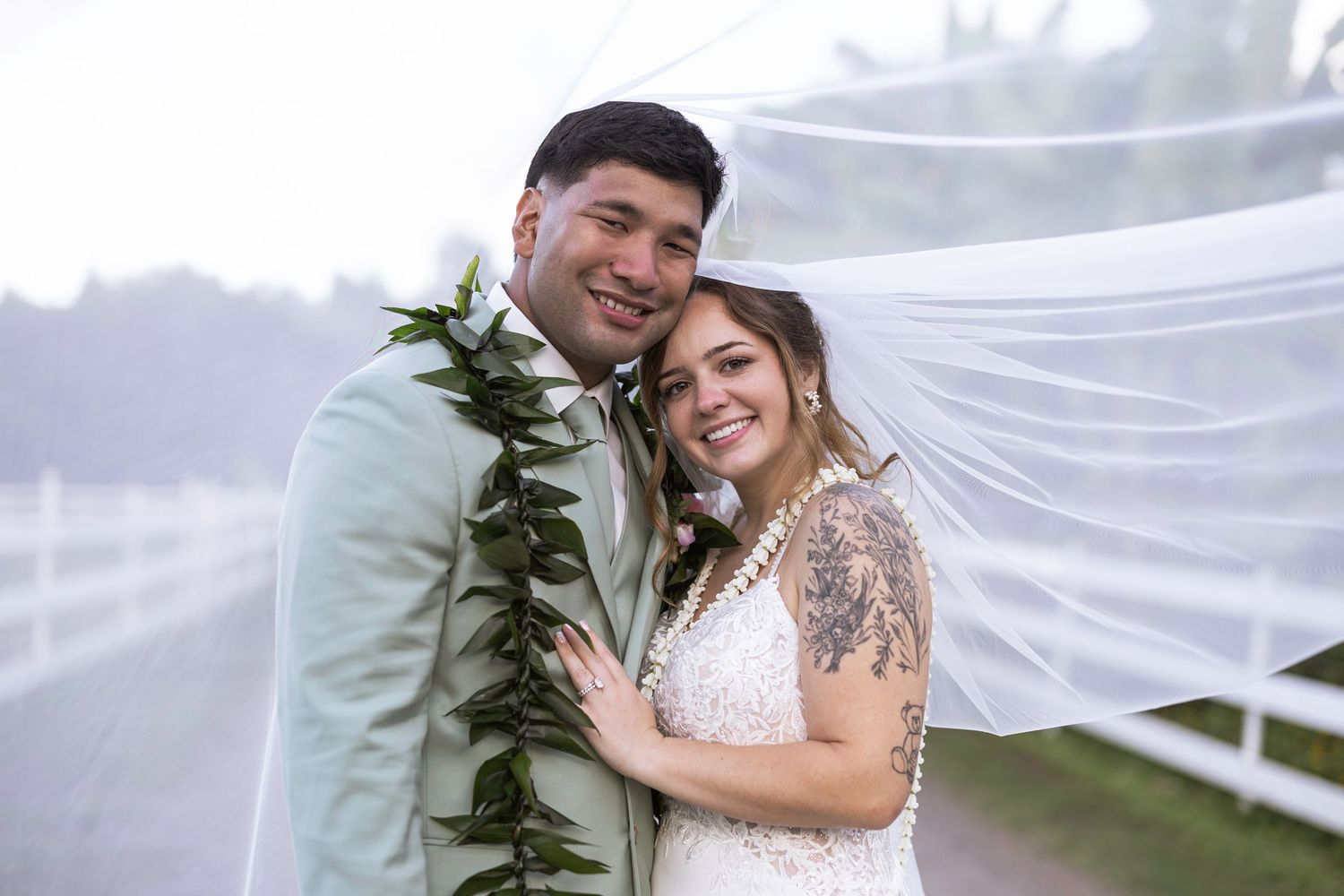 A couple embraces during their Hawaiian wedding ceremony with a maile lei and flowing veil in the misty background.