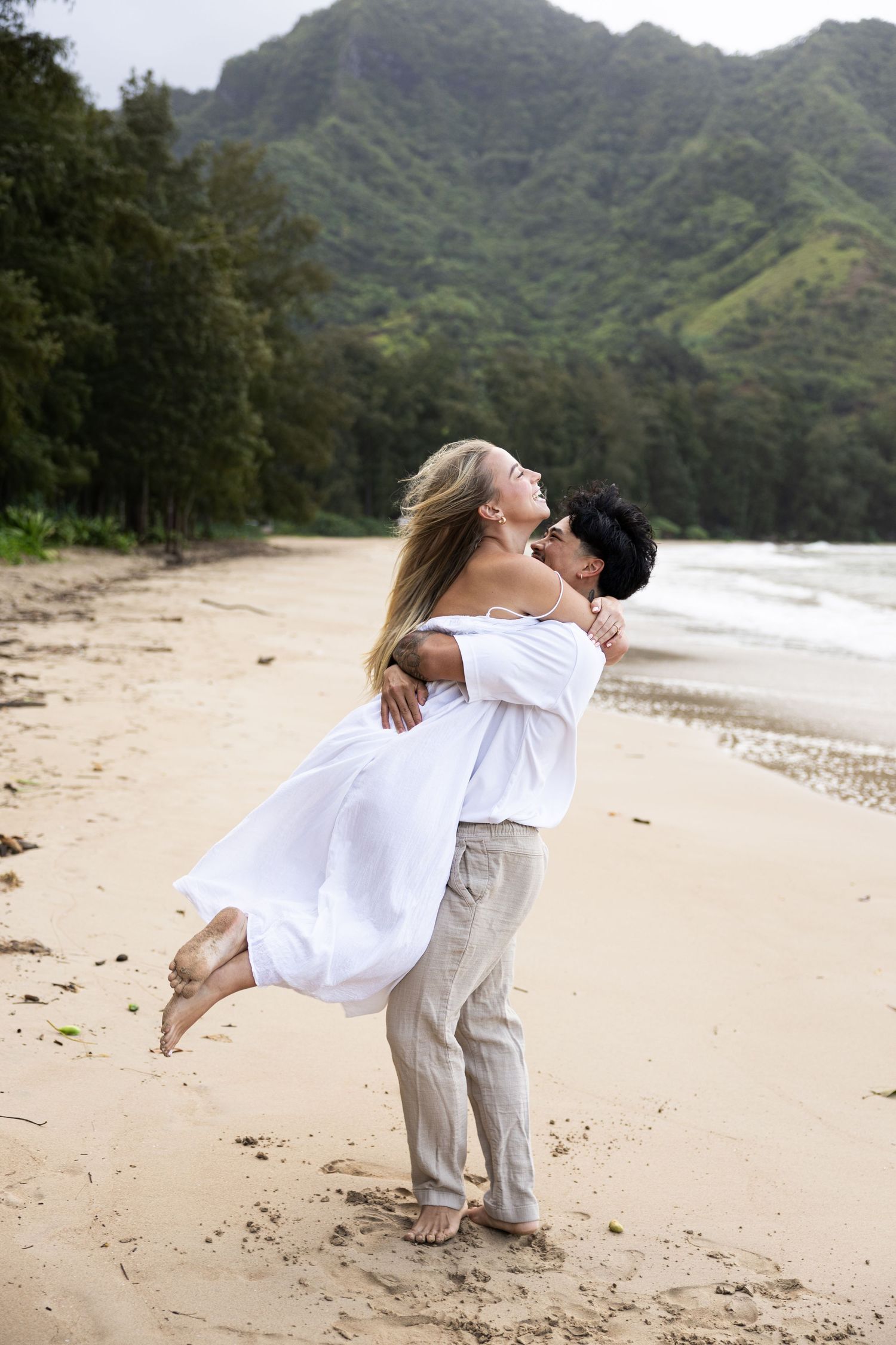A couple shares a joyful moment as one lifts the other on a tropical beach with mountains behind them.