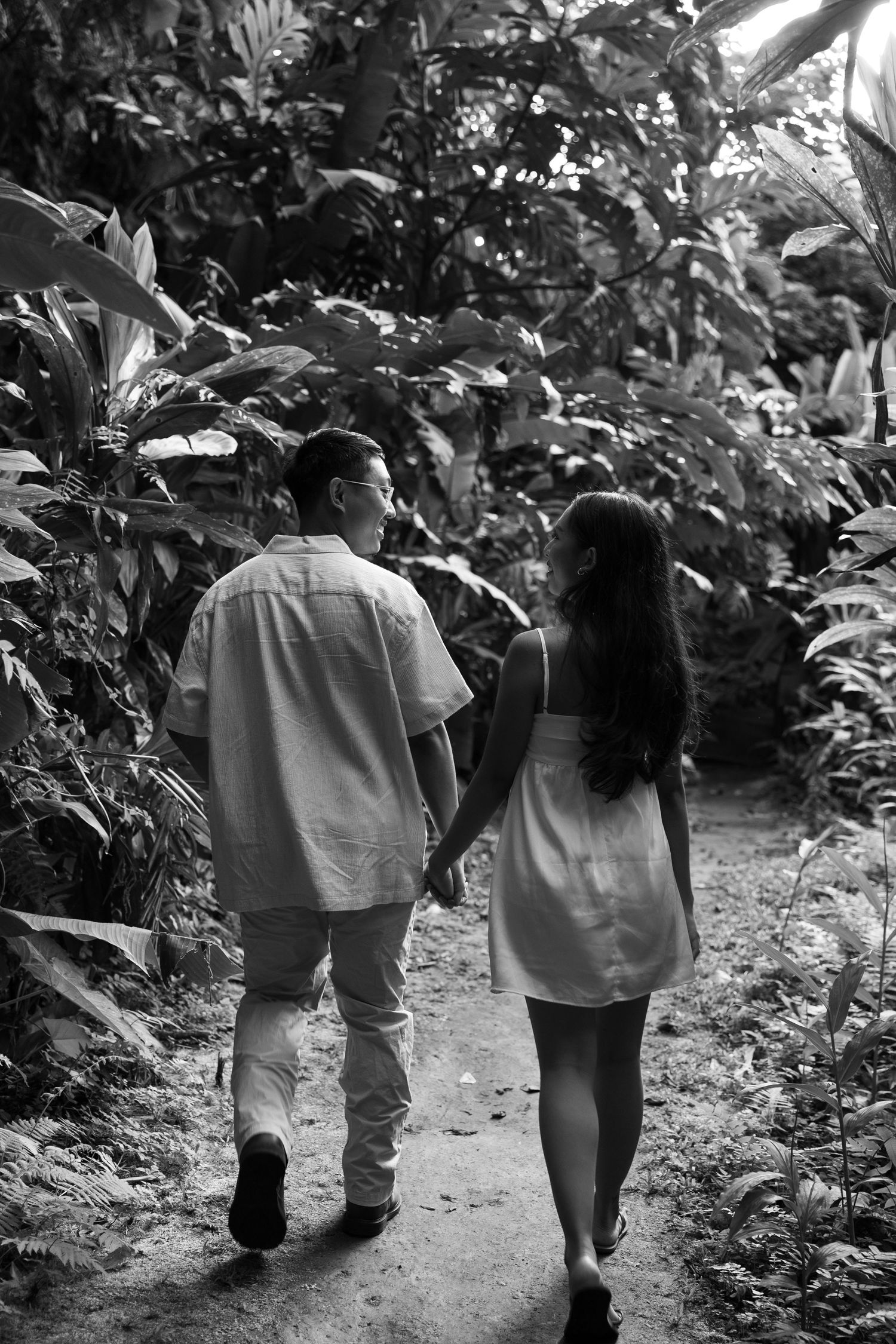 A couple walks hand in hand down a path surrounded by tropical foliage in black and white.