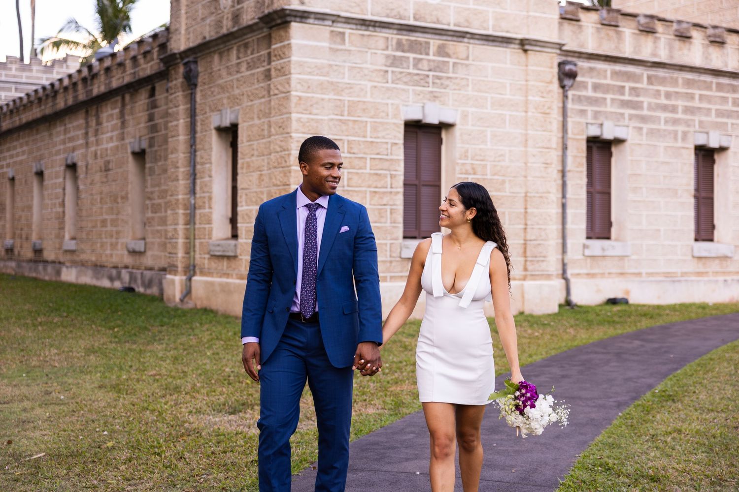 A couple walks together on grounds of a historic stone building, the bride holding a small bouquet.