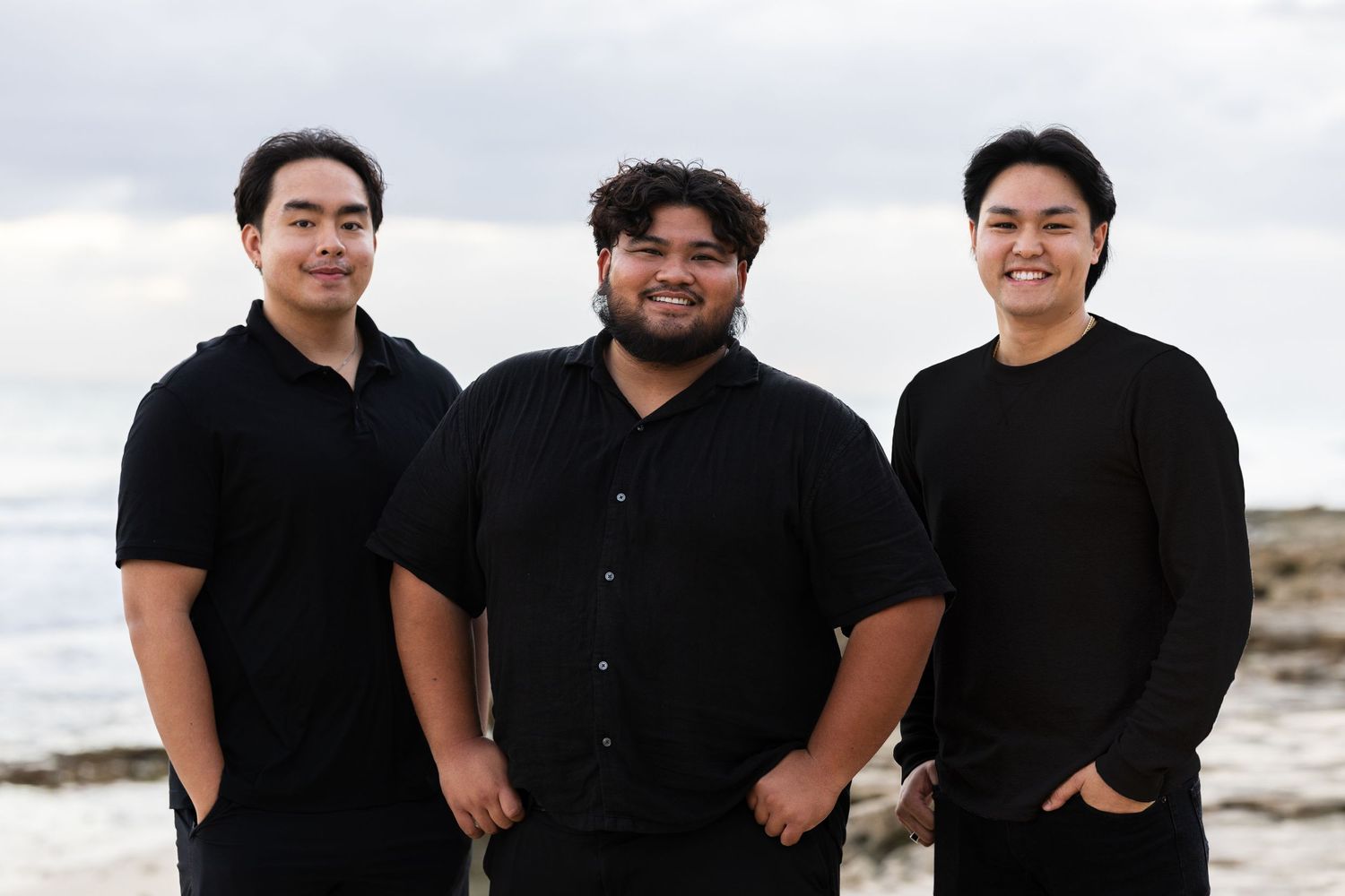 A group of people wearing black outfits pose together for a casual photo shoot at a beach location.