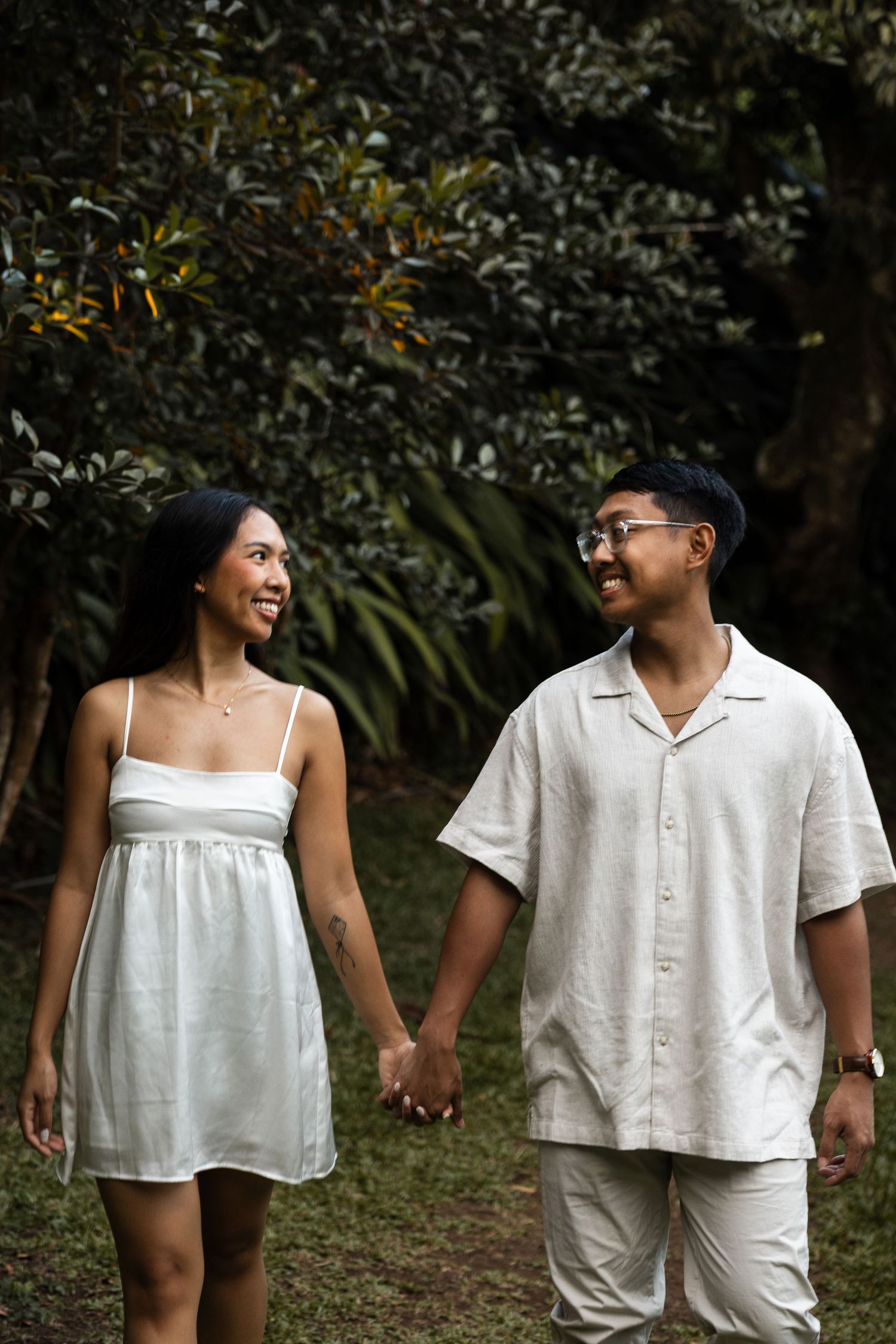 A couple in white clothing holds hands while walking through a lush garden path.