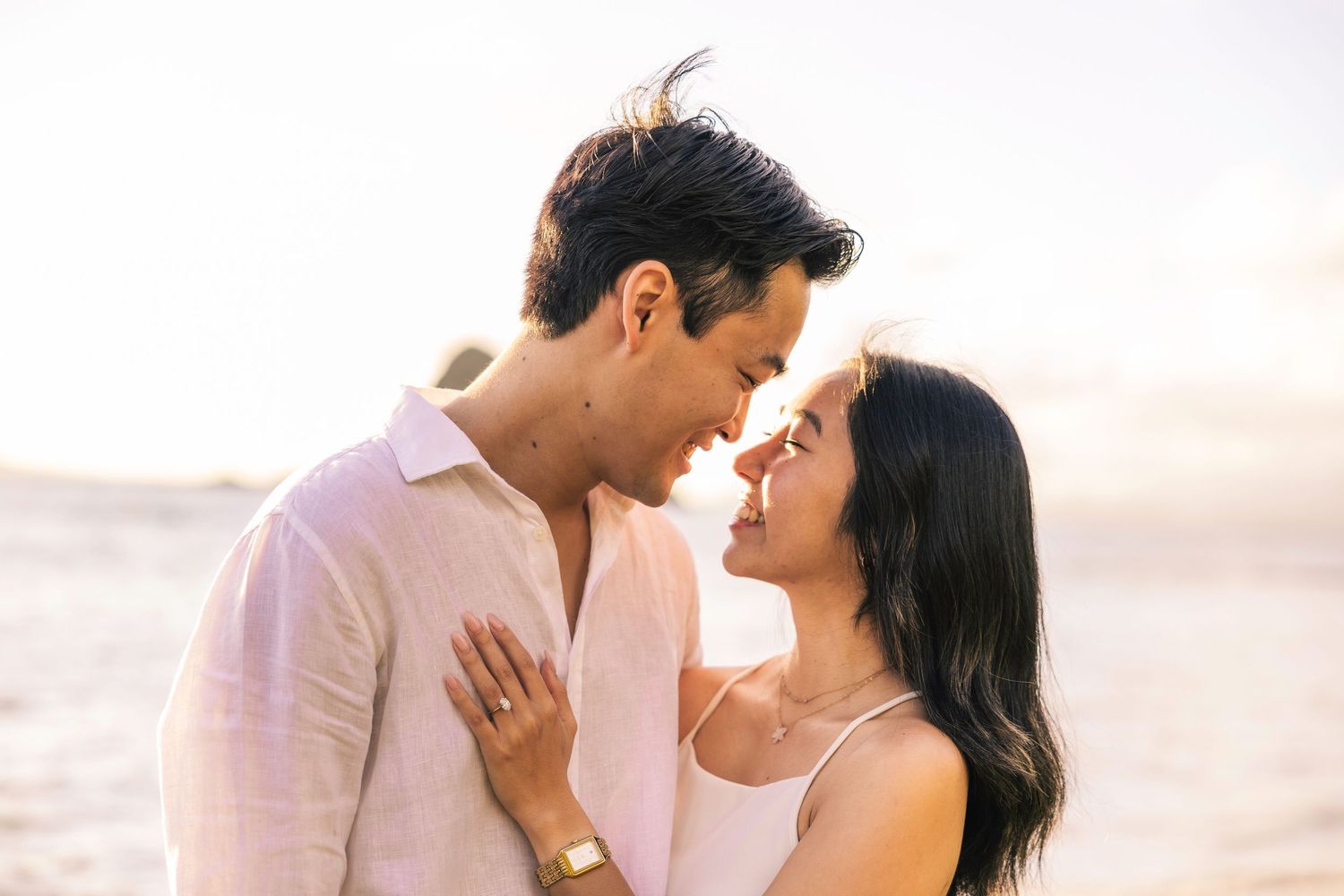 A romantic couple shares tender moments at sunset on the beach in white and pink outfits.