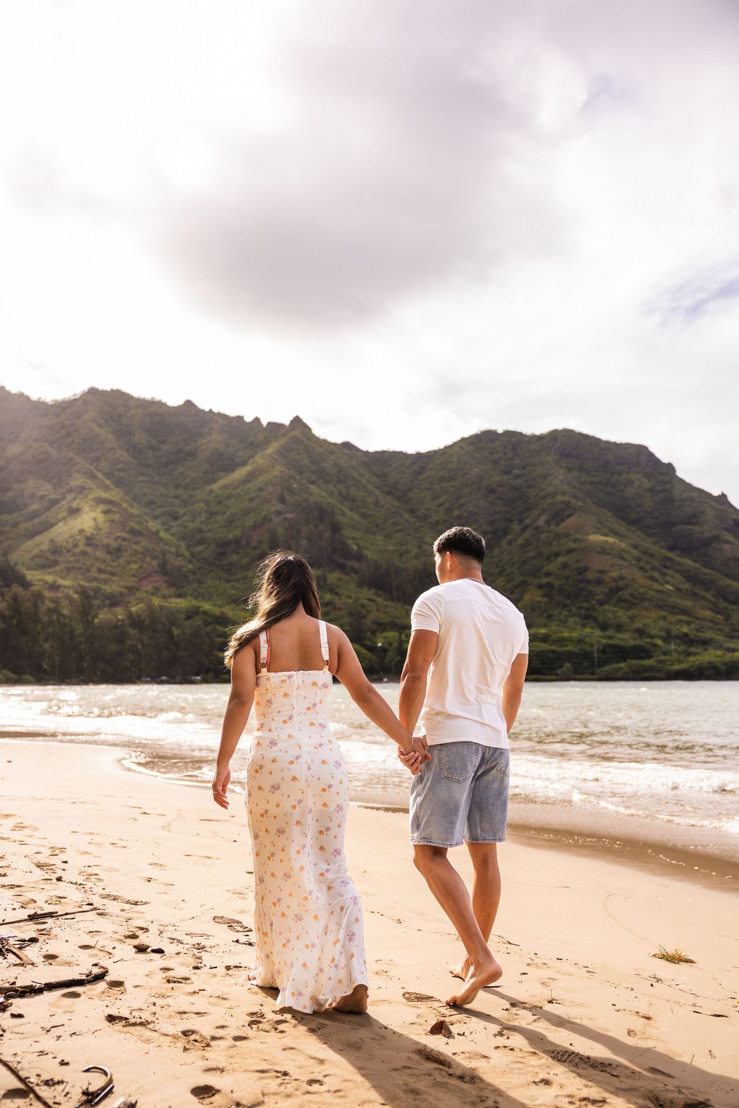 A couple walking hand in hand on a tropical beach with dramatic mountain cliffs in the background.