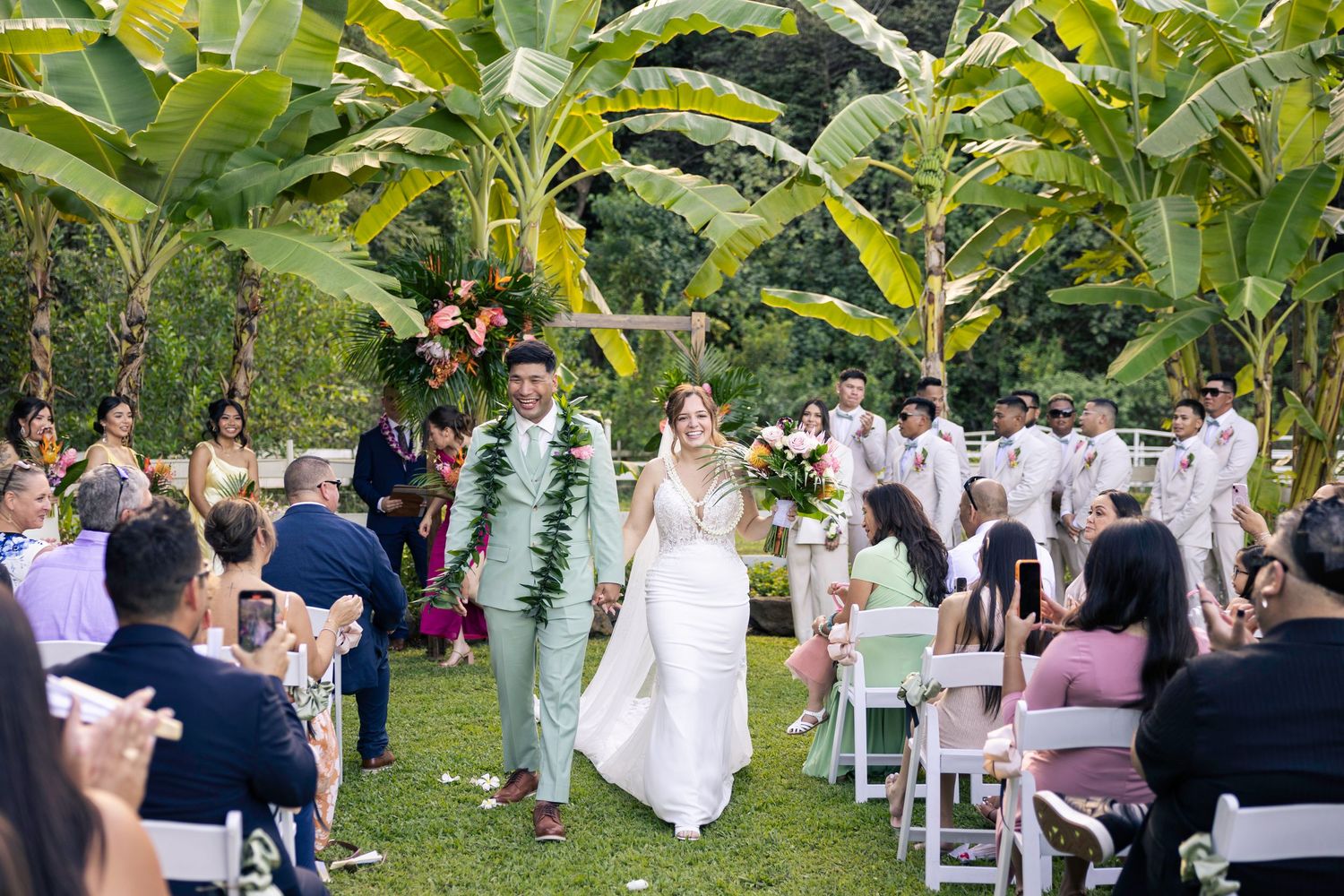 Wedding guests seated in white chairs watch a ceremony procession under tropical foliage.