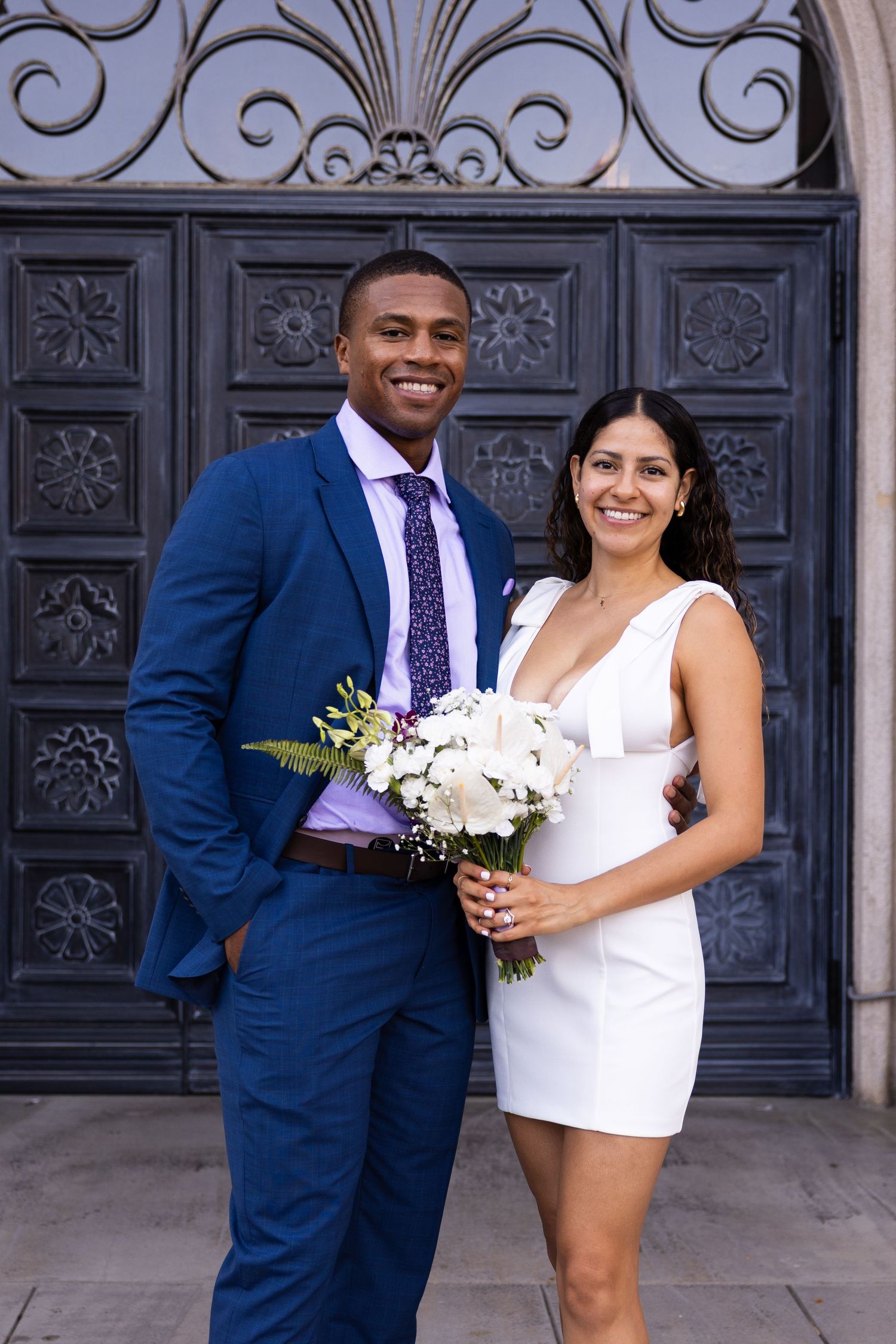 A couple poses together in wedding attire, with the bride in white and groom in navy suit against an ornate door background.