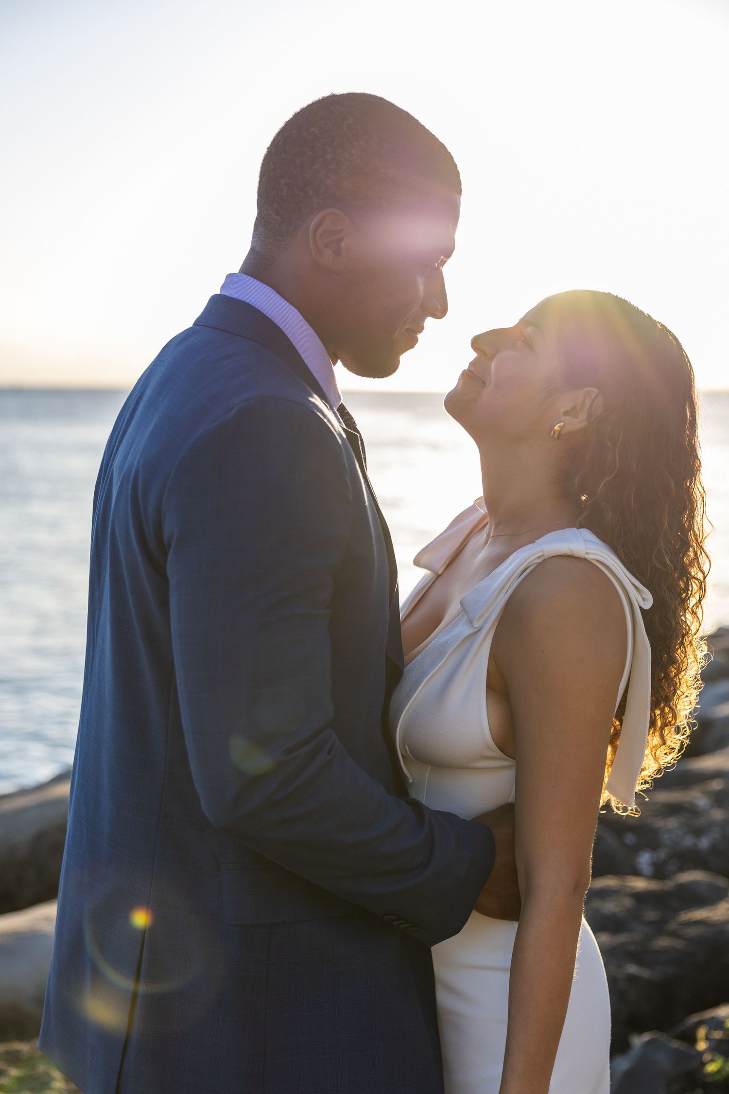 Backlit couple in formal attire sharing intimate moment by ocean at sunset.
