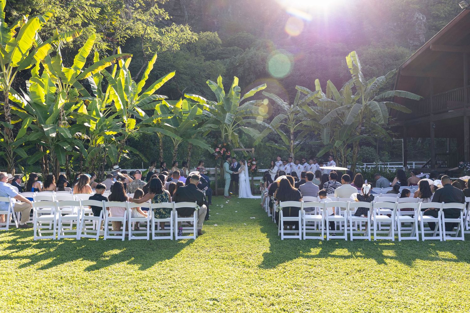 Sun flares illuminate empty white chairs set up for an outdoor wedding ceremony.