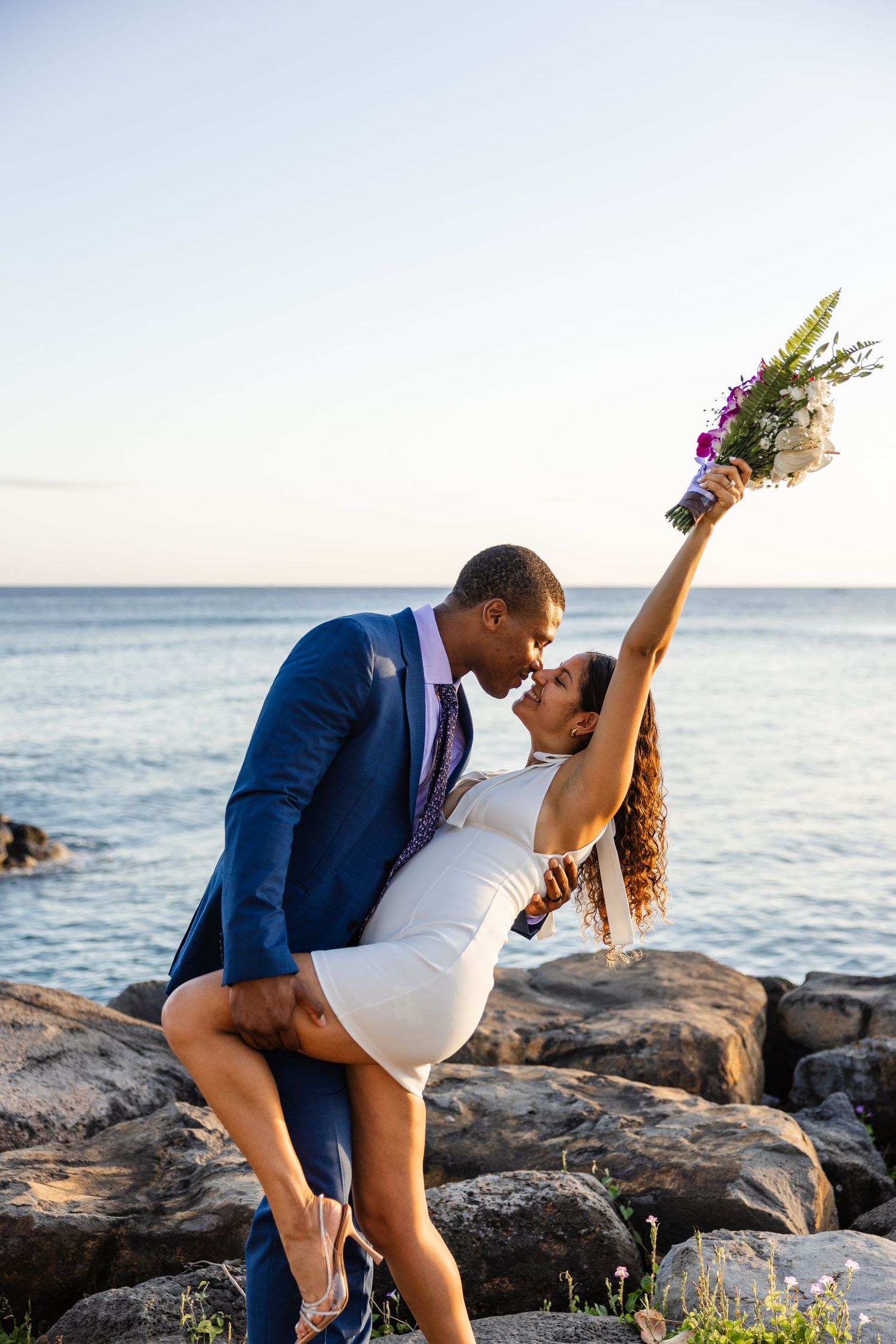Playful wedding pose with bride holding bouquet up while dipping on coastal rocks.