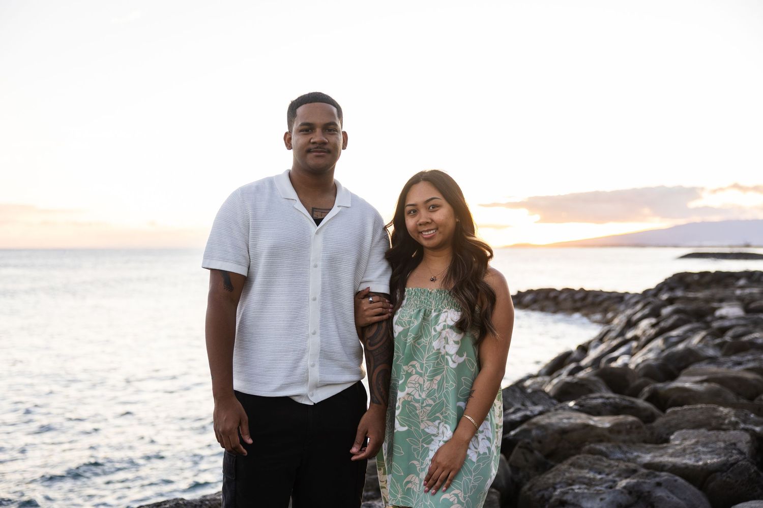 Couple stands together on rocky beach at sunset wearing white and mint green outfits for romantic photoshoot.