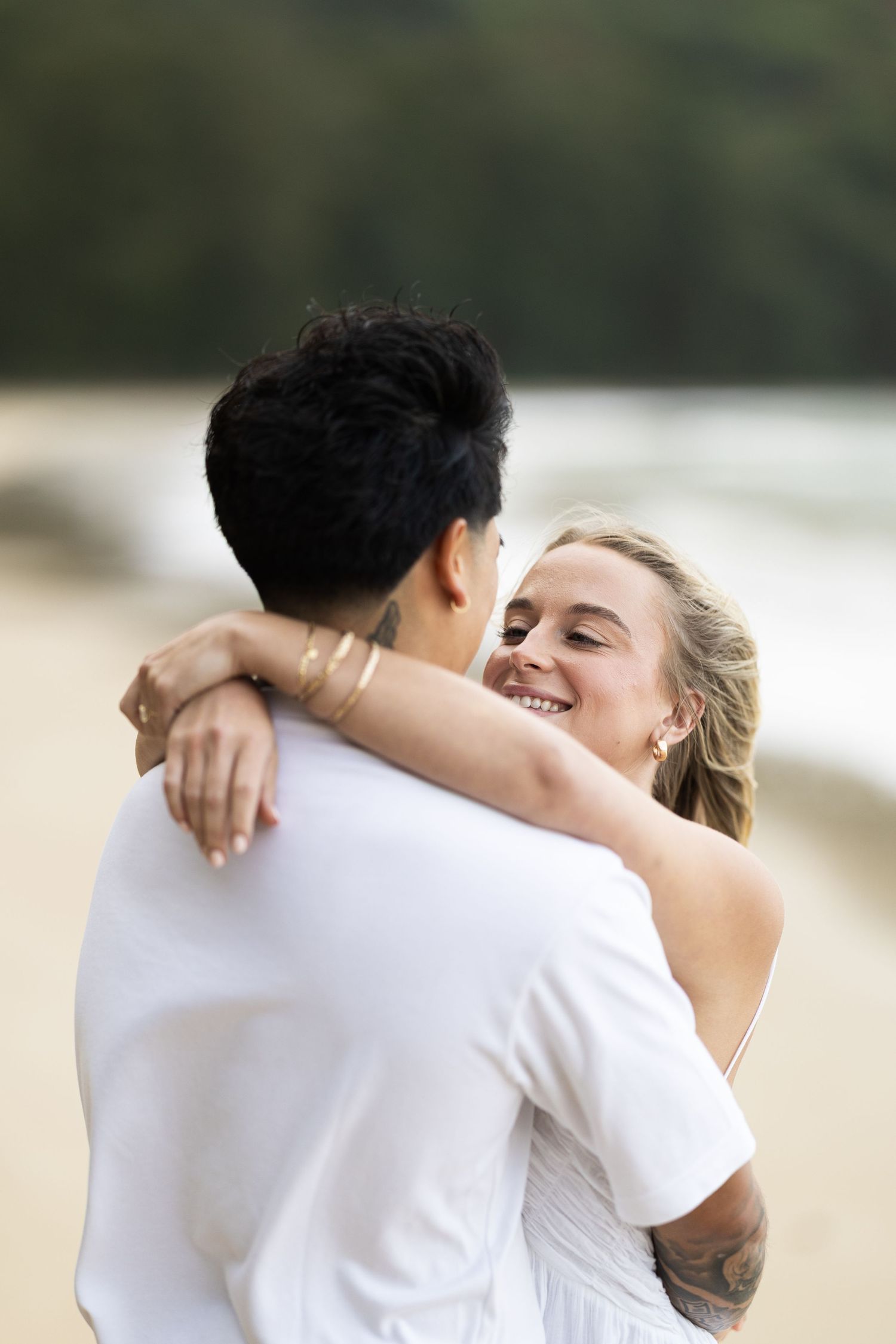 A couple embraces in white outfits on a beach with blurred background.
