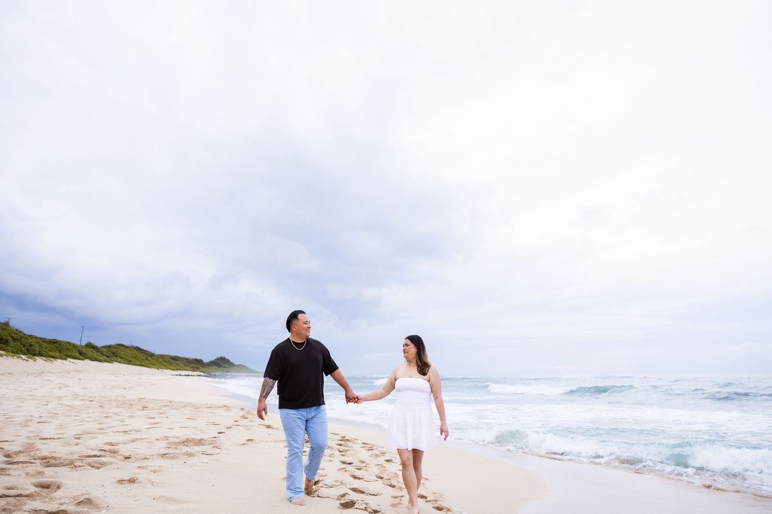 Two people walk hand in hand along a sandy beach with ocean waves and cloudy skies in the background.