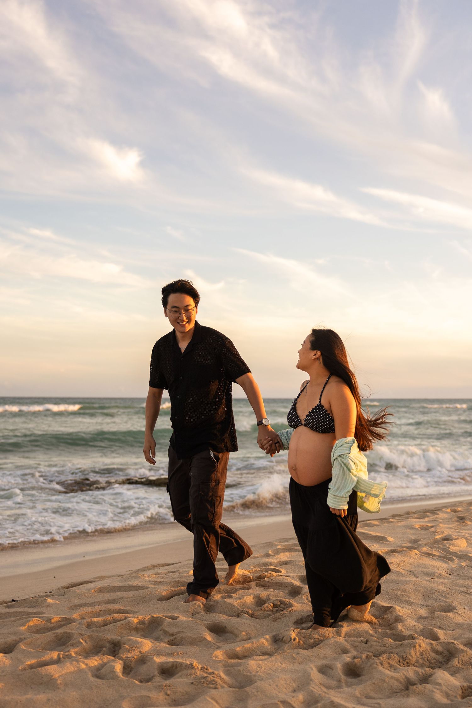 Two people run playfully along a sandy beach at sunset with waves crashing in the background.