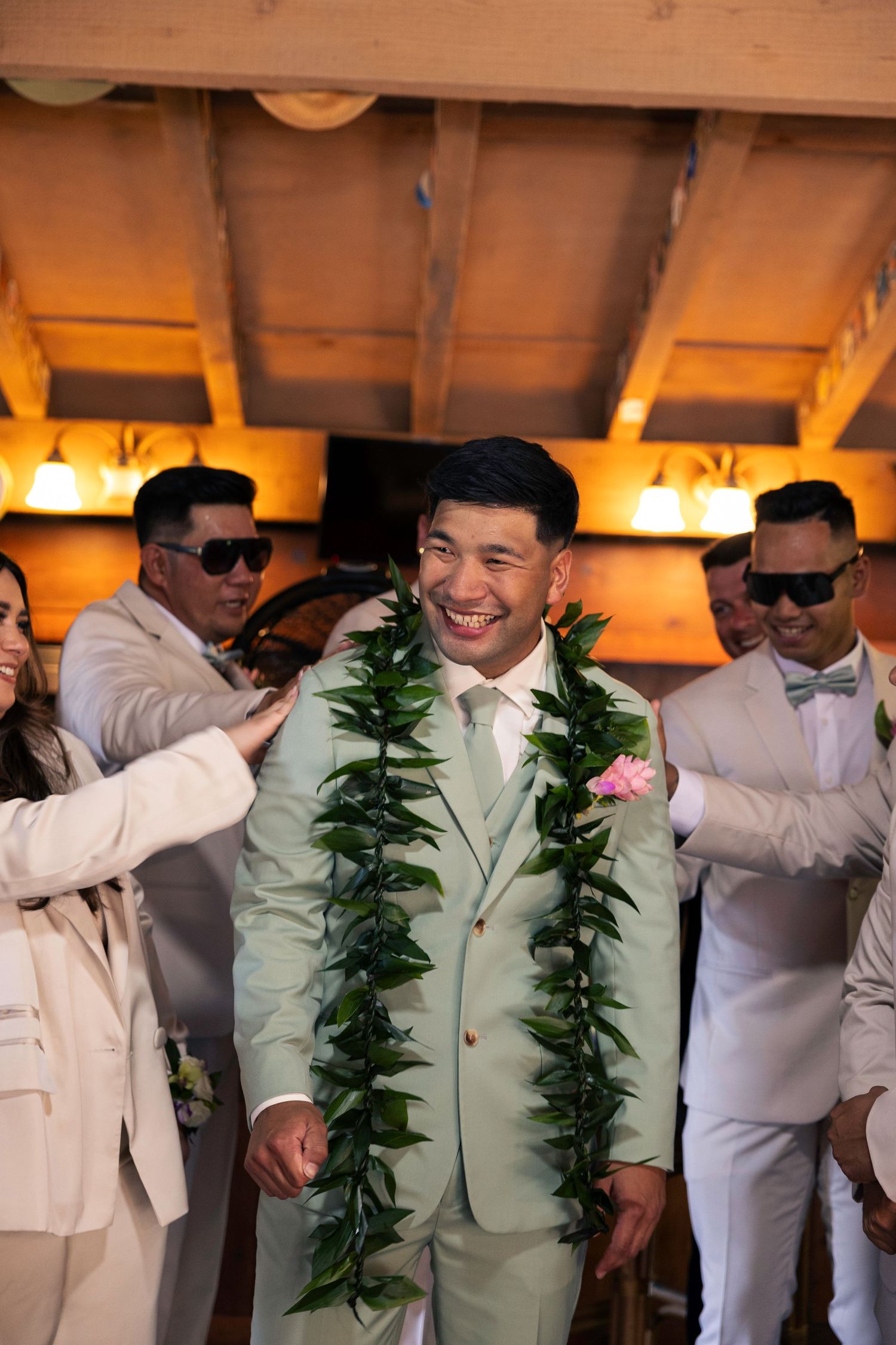 A joyful wedding celebration with the groom wearing a light suit and traditional maile lei surrounded by guests in white attire.