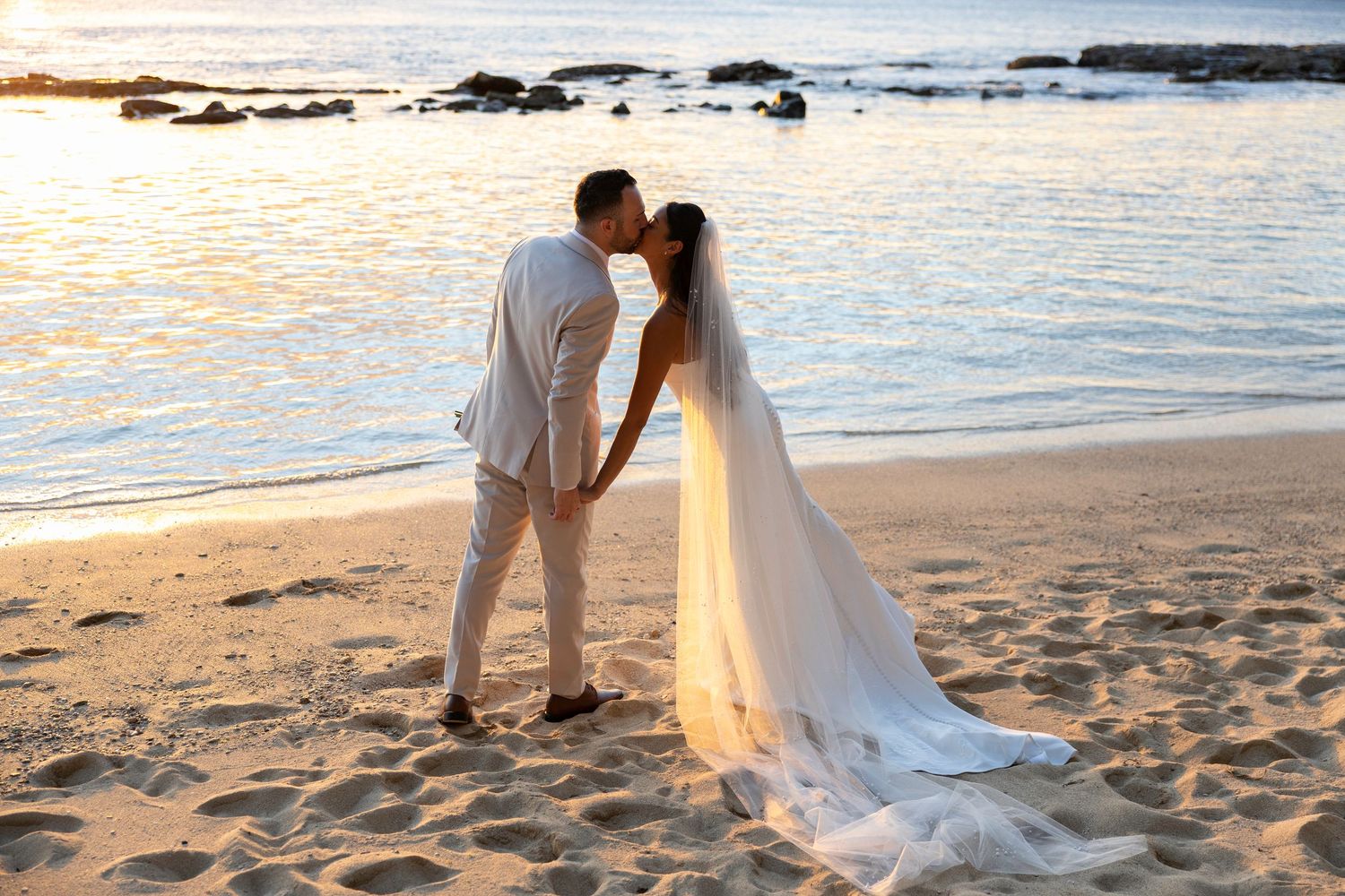 A romantic beach wedding scene at sunset with a couple kissing on golden sand by the ocean.