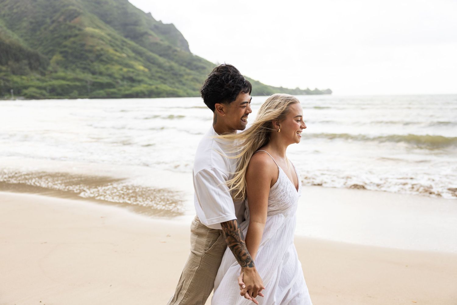 A couple walks hand in hand along a scenic Hawaiian beach with mountains in the background.