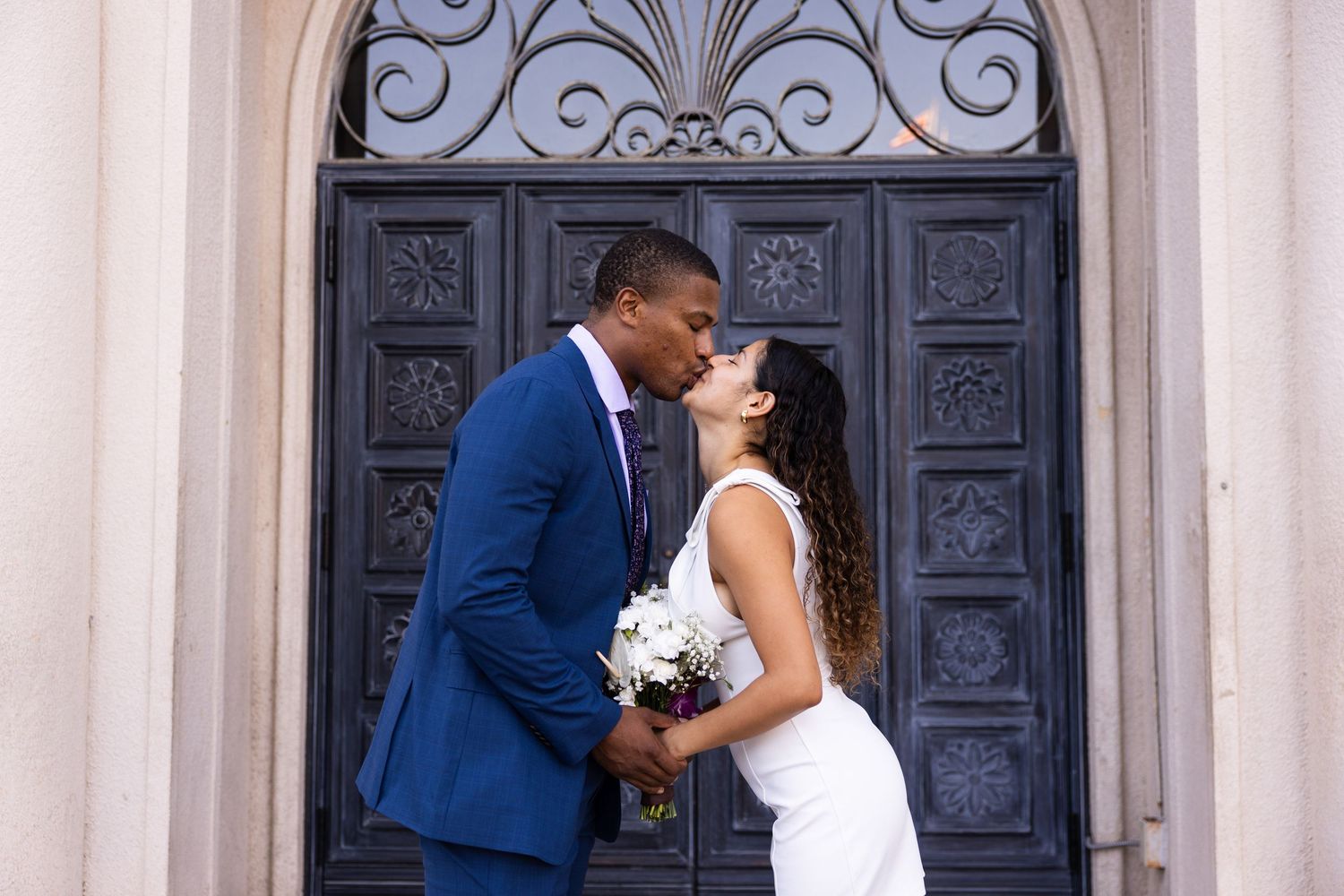 Newlyweds share a romantic kiss in front of an ornate dark door at a historic building entrance.