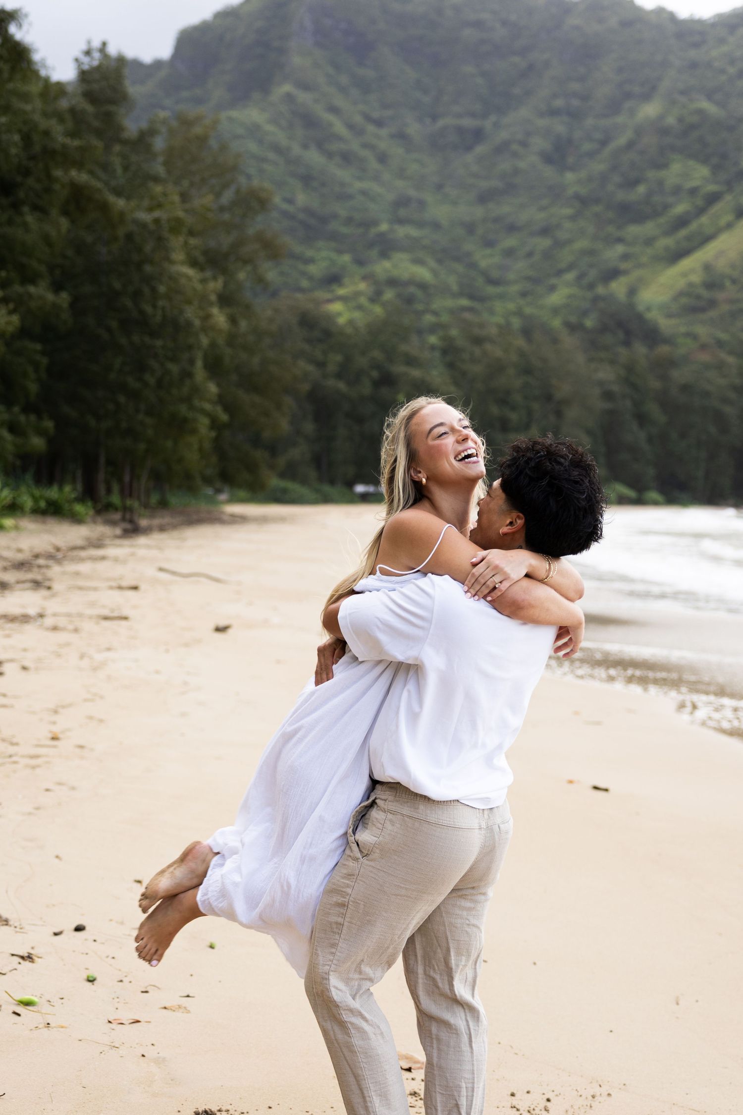 A couple in white clothing laughs and embraces joyfully on a scenic beach with mountains in the background.