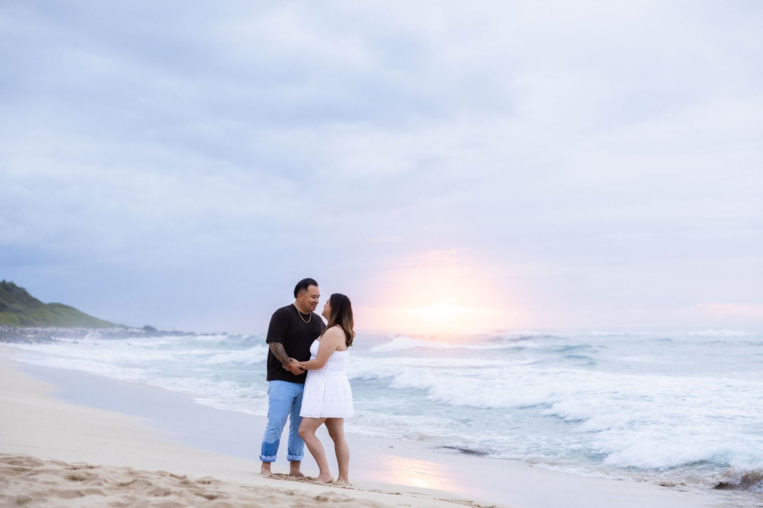 A romantic couple shares a tender moment on a misty beach at sunset with waves crashing in the background.