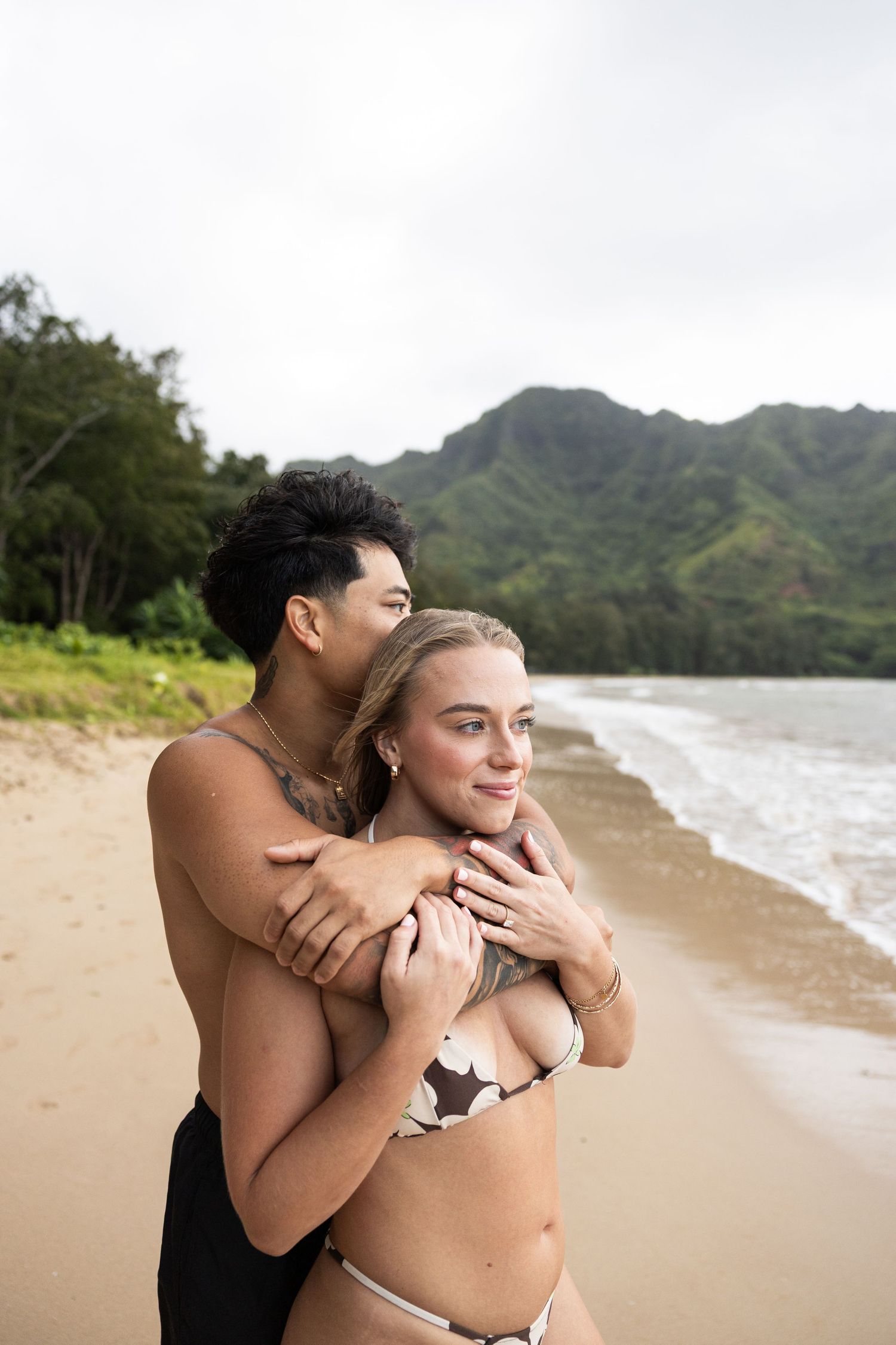 Couple embracing on a scenic beach with lush mountains in Hawaii.