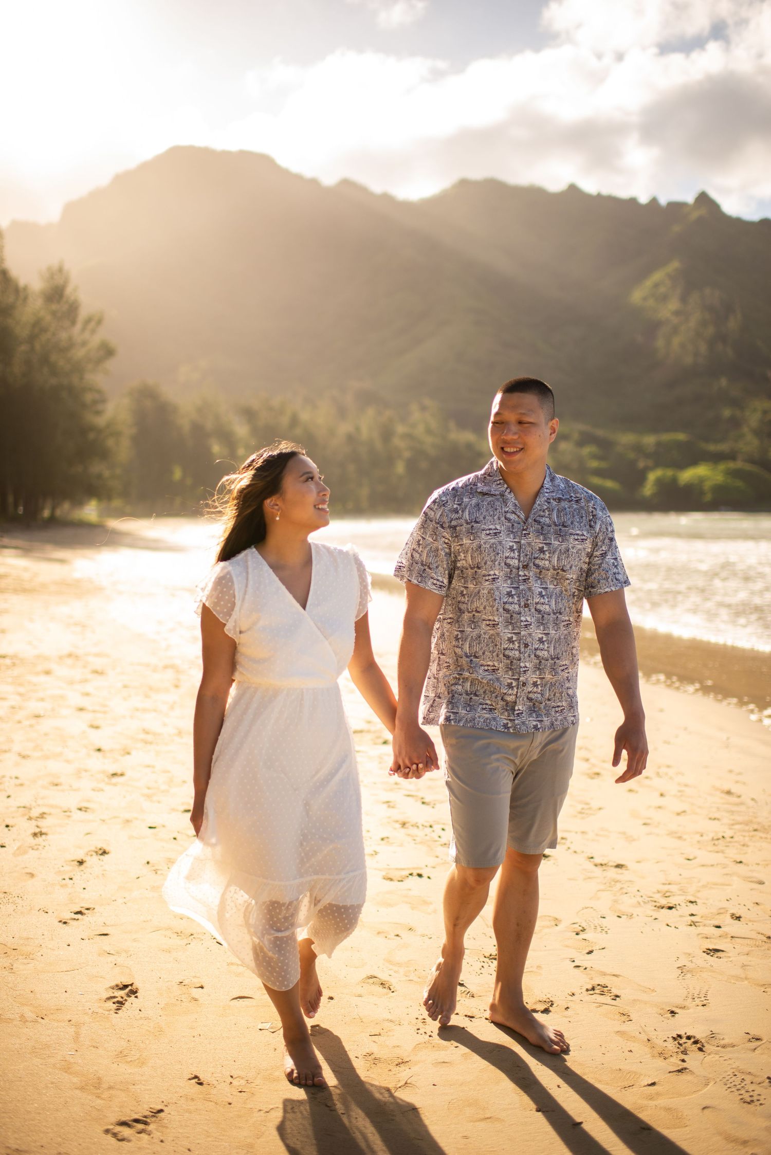 A couple walking hand in hand along a scenic beach at sunset with mountains in the background.