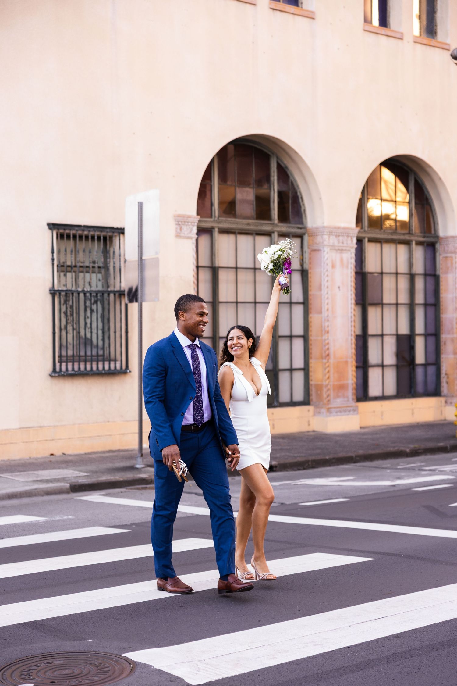 A stylish couple poses on a crosswalk in front of an arched building facade.