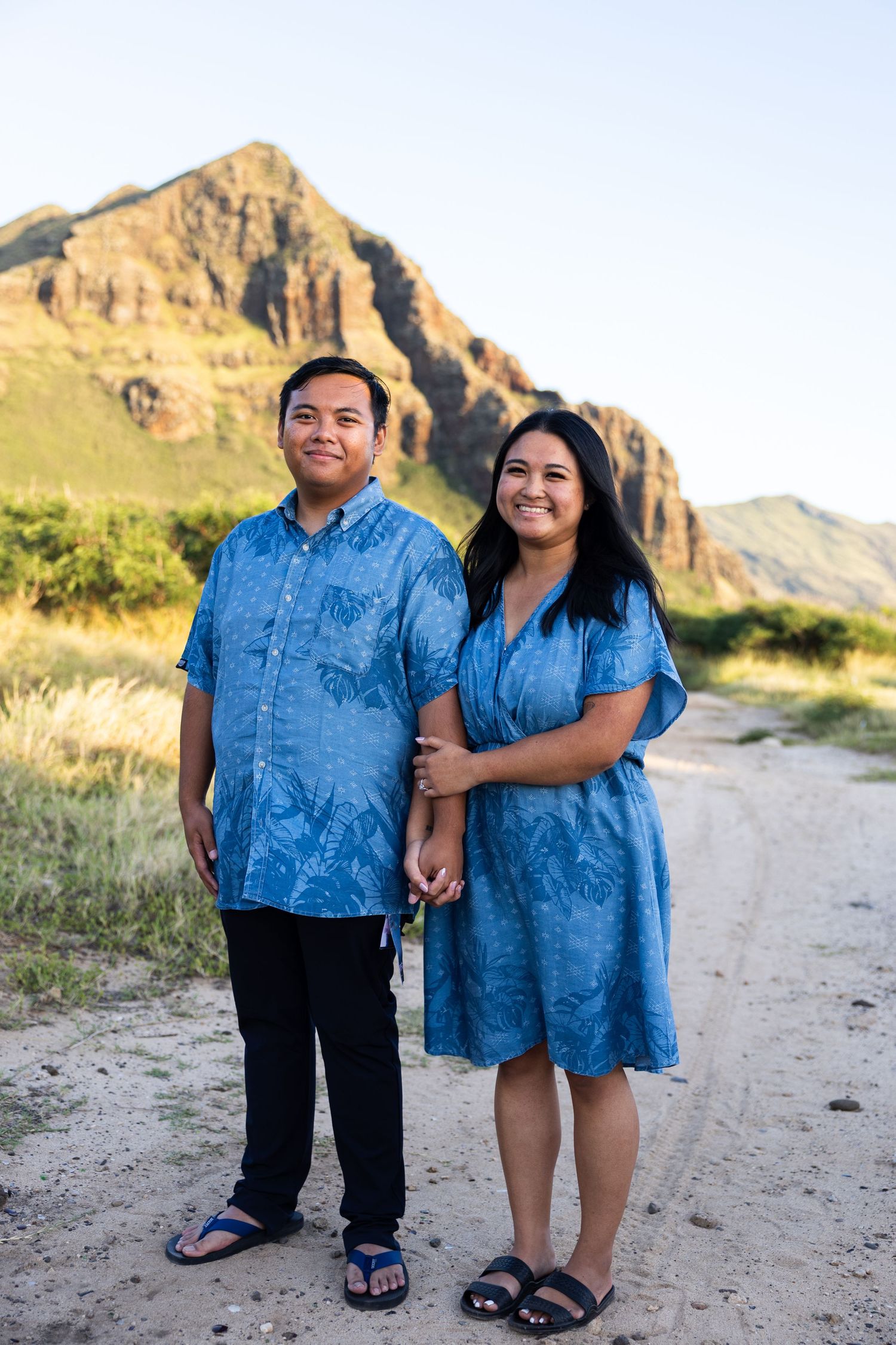 A couple wearing matching blue outfits poses together on a desert trail with rugged mountains in the background.