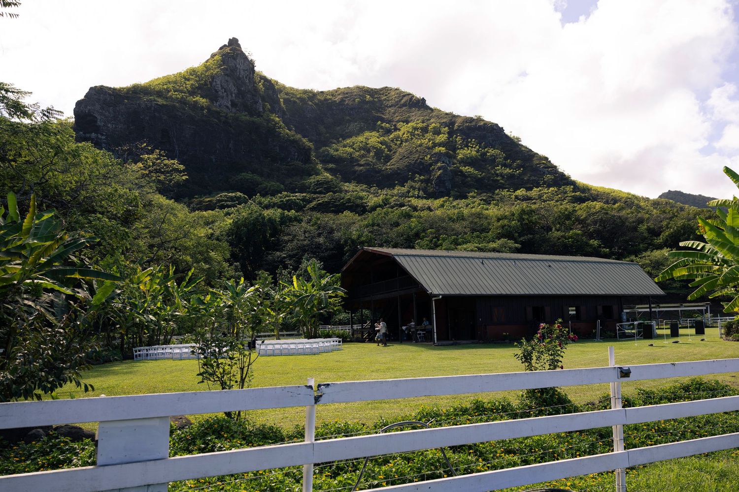 Black barn building with white fence in front of dramatic mountain landscape in tropical setting.