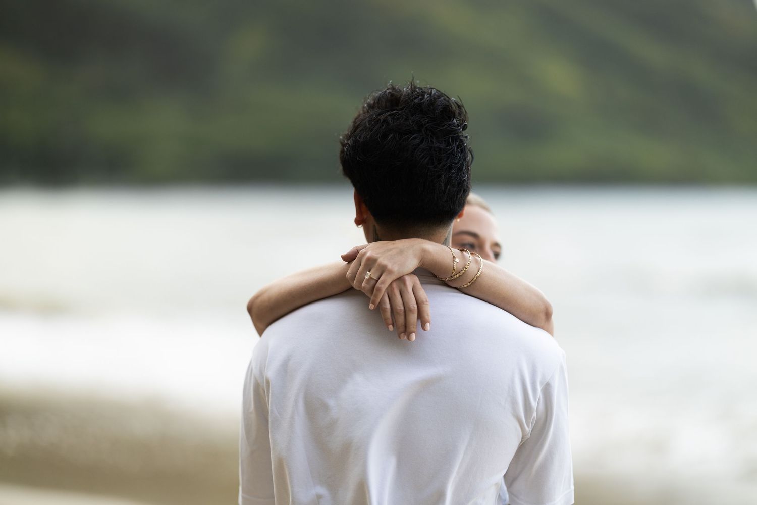 A person embraces another from behind while overlooking a serene beach landscape.