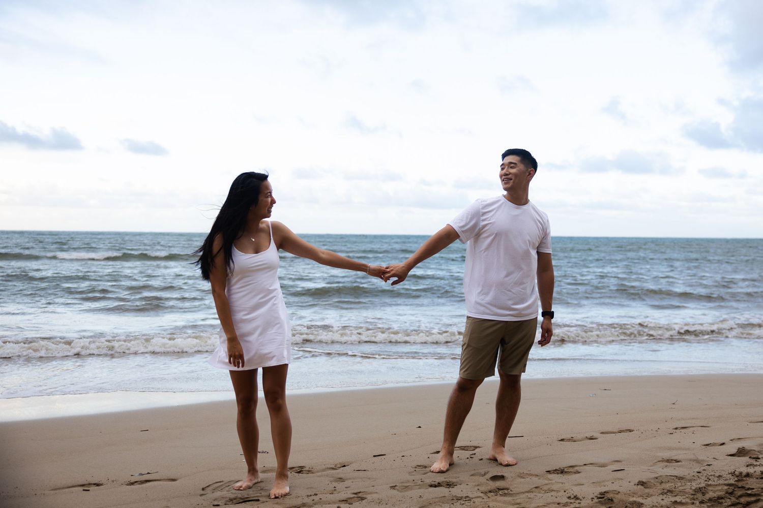 Two people walking together on a sandy beach at sunset holding hands with ocean waves in the background.