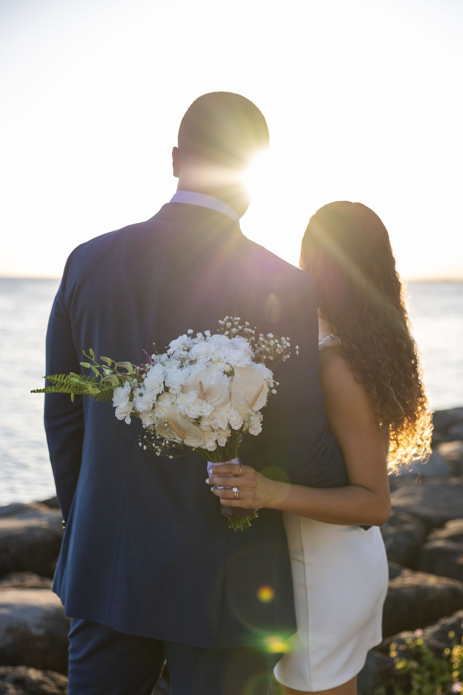 Silhouetted couple stands by ocean at sunset with bride holding white bouquet.