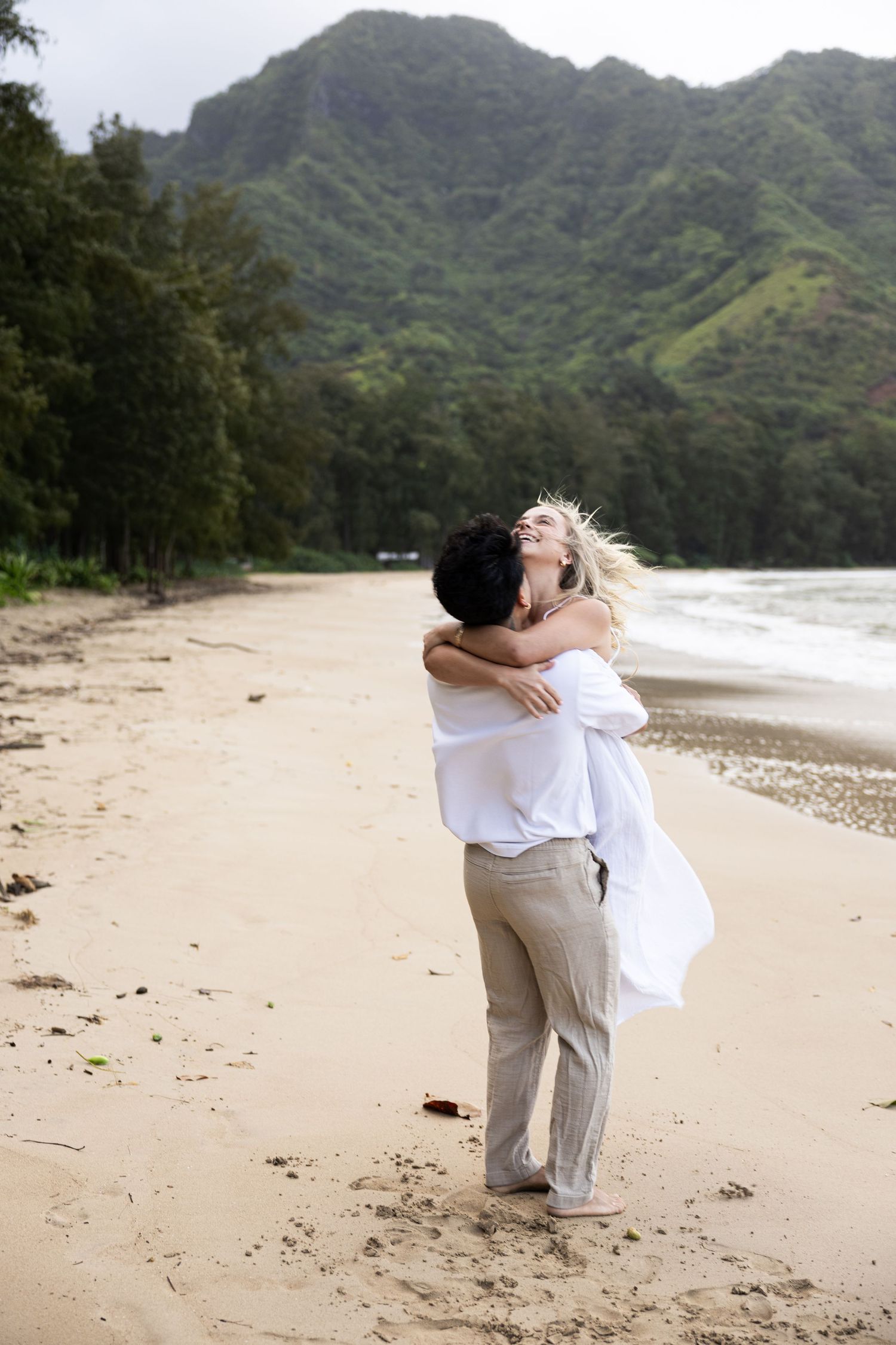 Two lovers embrace in a romantic hug on a scenic Hawaiian beach with lush mountains in the background.