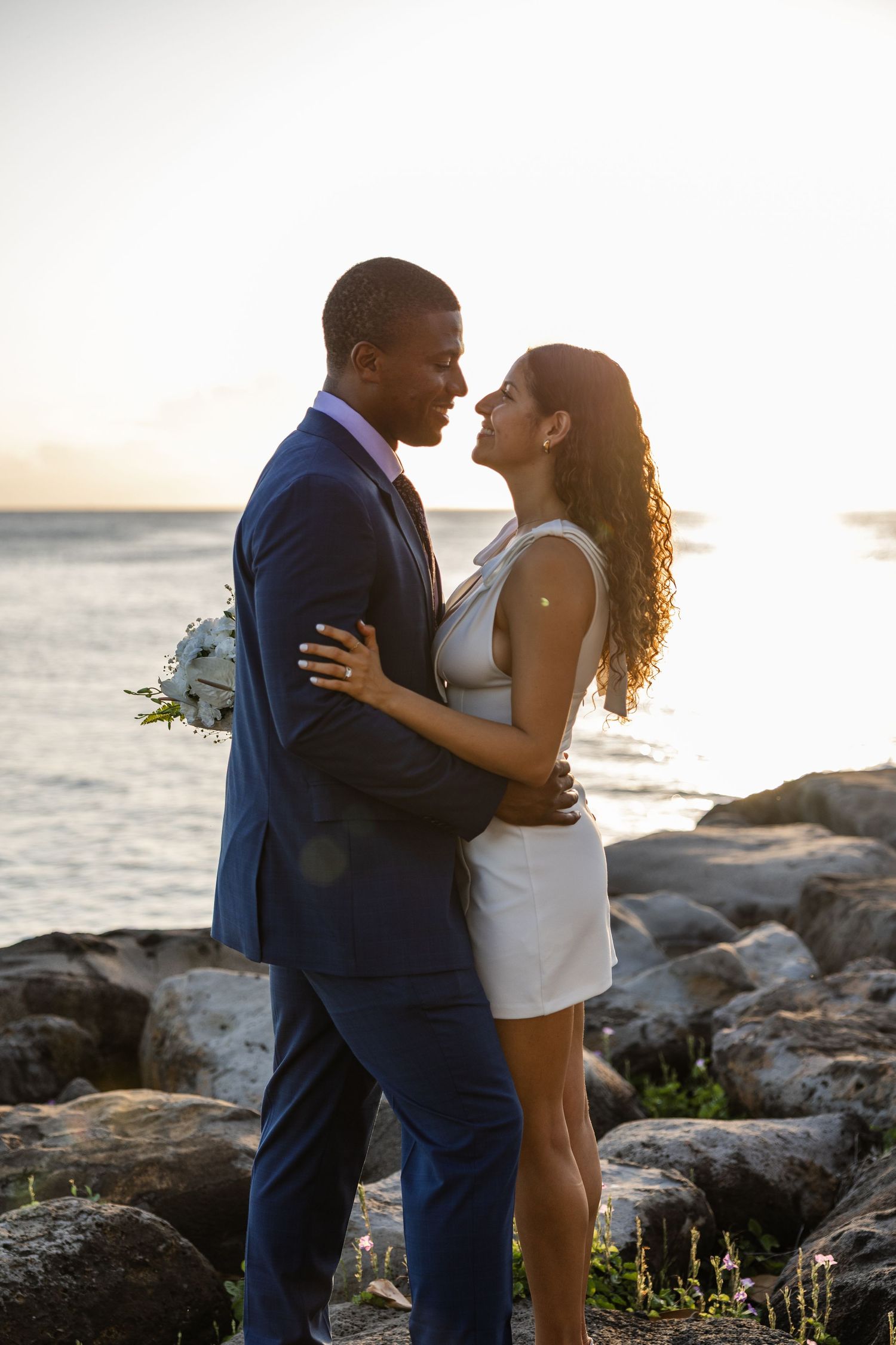 Couple embracing on rocky beach during sunset with ocean in background.