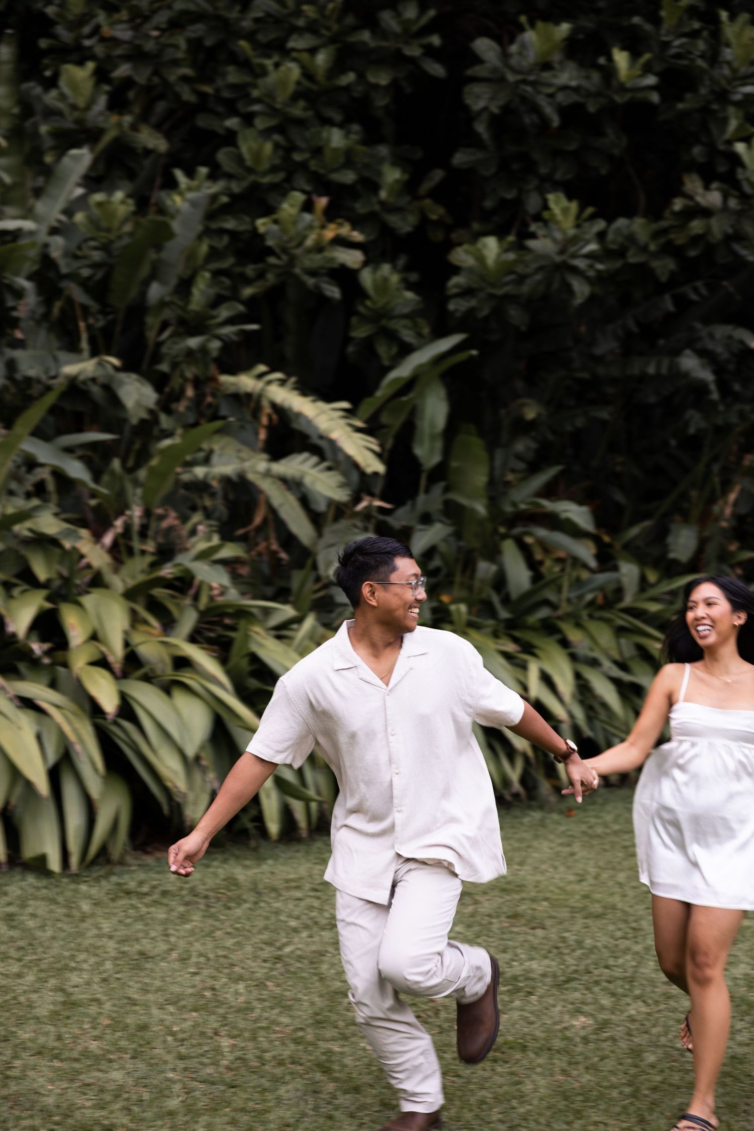 A playful couple in matching white outfits runs through a tropical garden landscape.