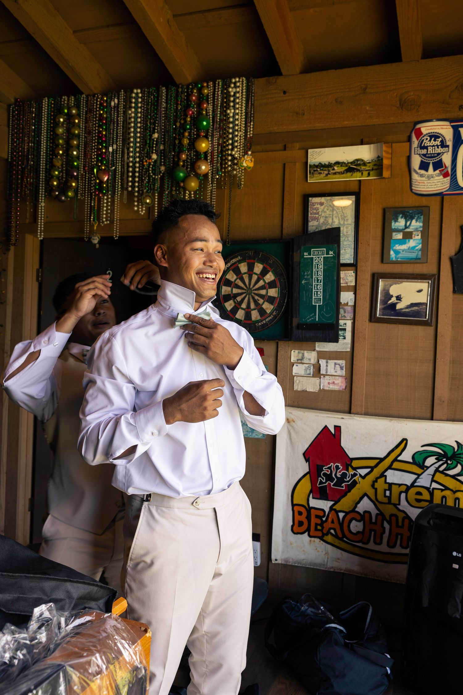 A person in a white outfit dances and poses playfully inside a rustic wooden room with a dartboard on the wall.