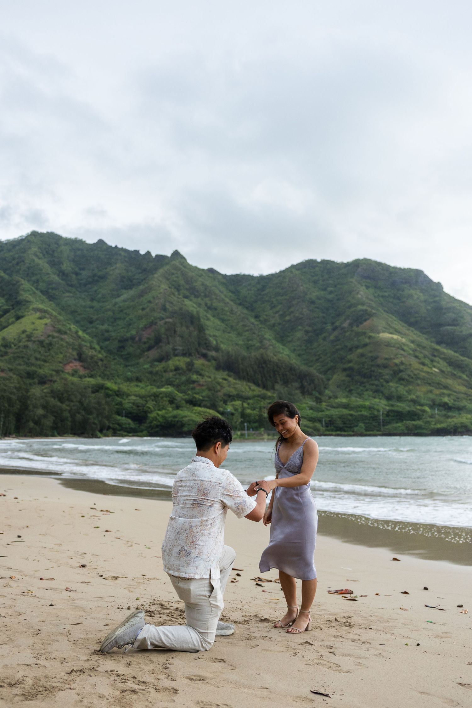 A couple shares a romantic moment during a proposal on a scenic beach with lush green mountains in the background.