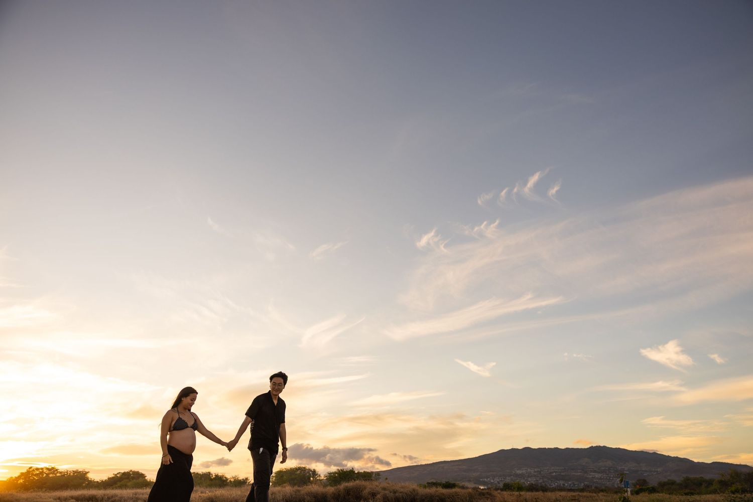 Silhouetted couple walking hand in hand along a beach at sunset with mountains in the background.