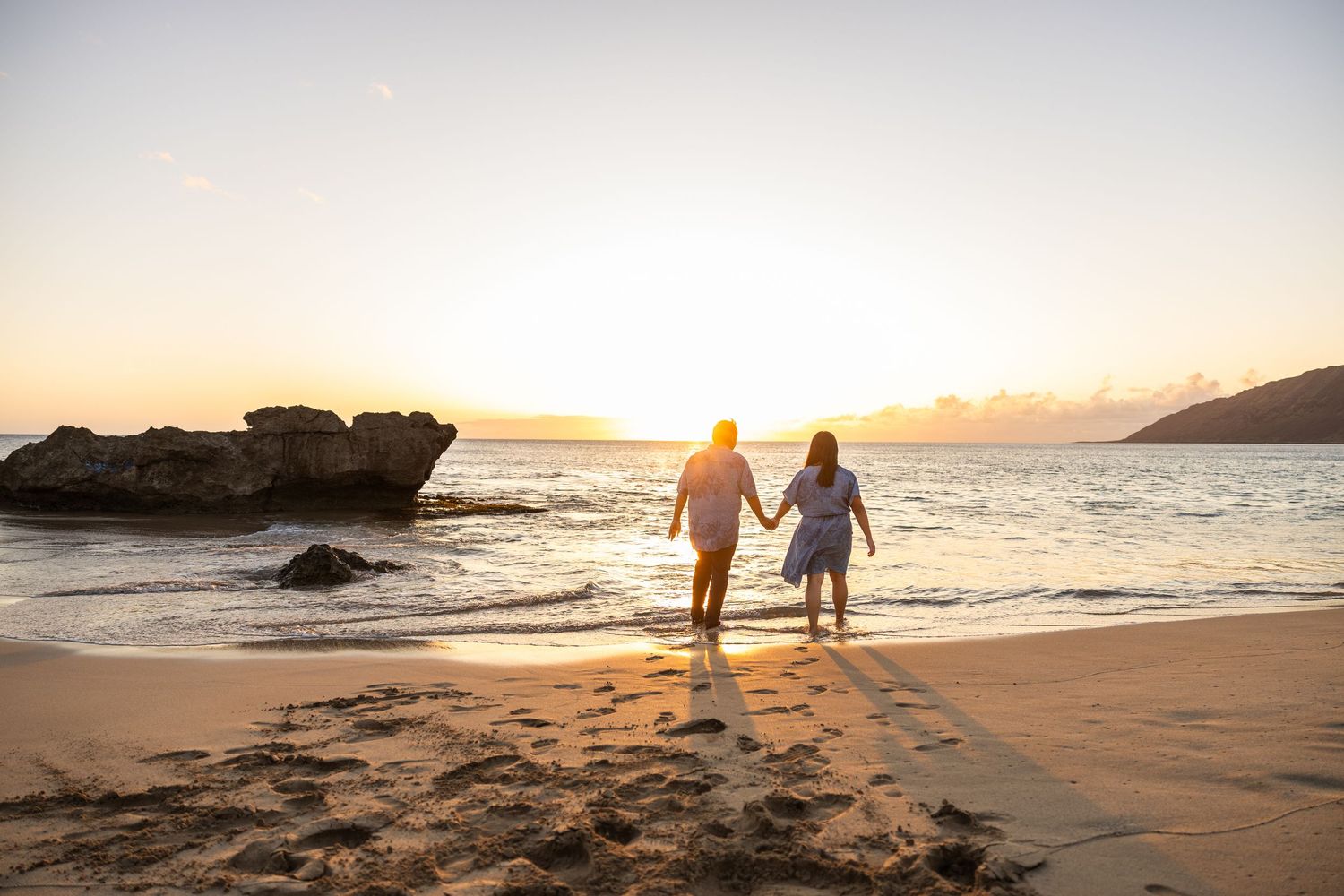 Two silhouettes walk along a sandy beach at sunset with golden light reflecting off the ocean waves.