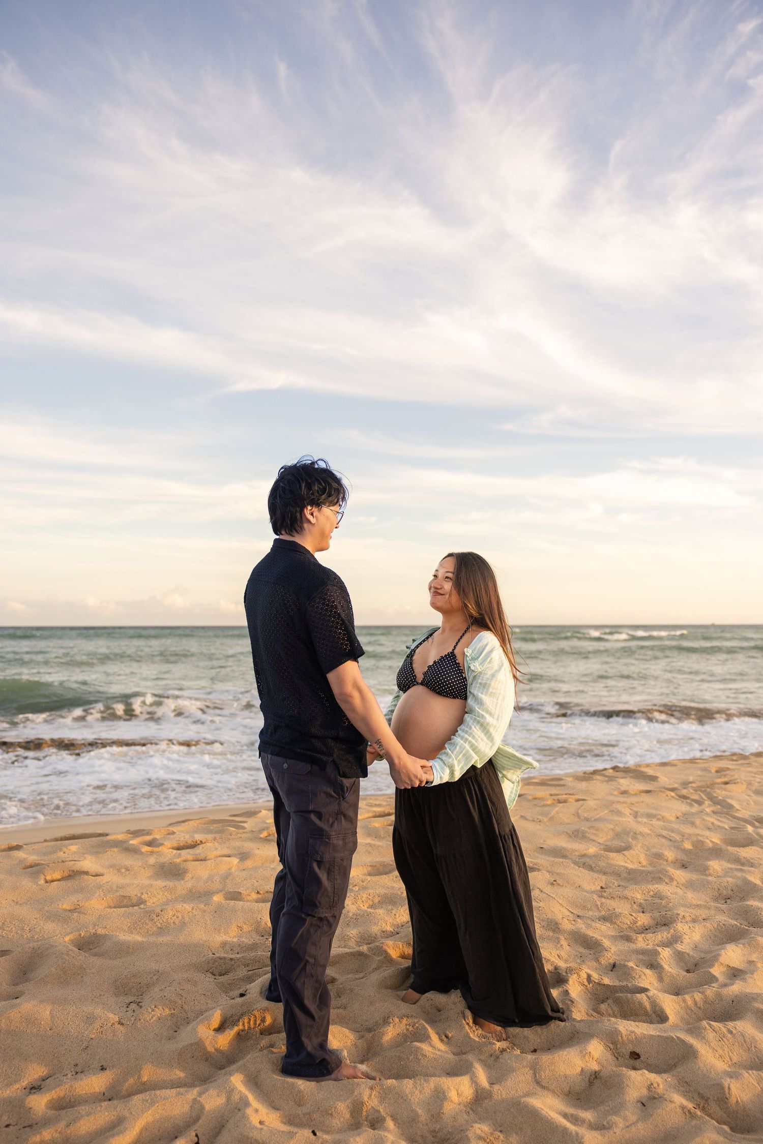 A couple stands together on a sandy beach at sunset with waves crashing behind them.