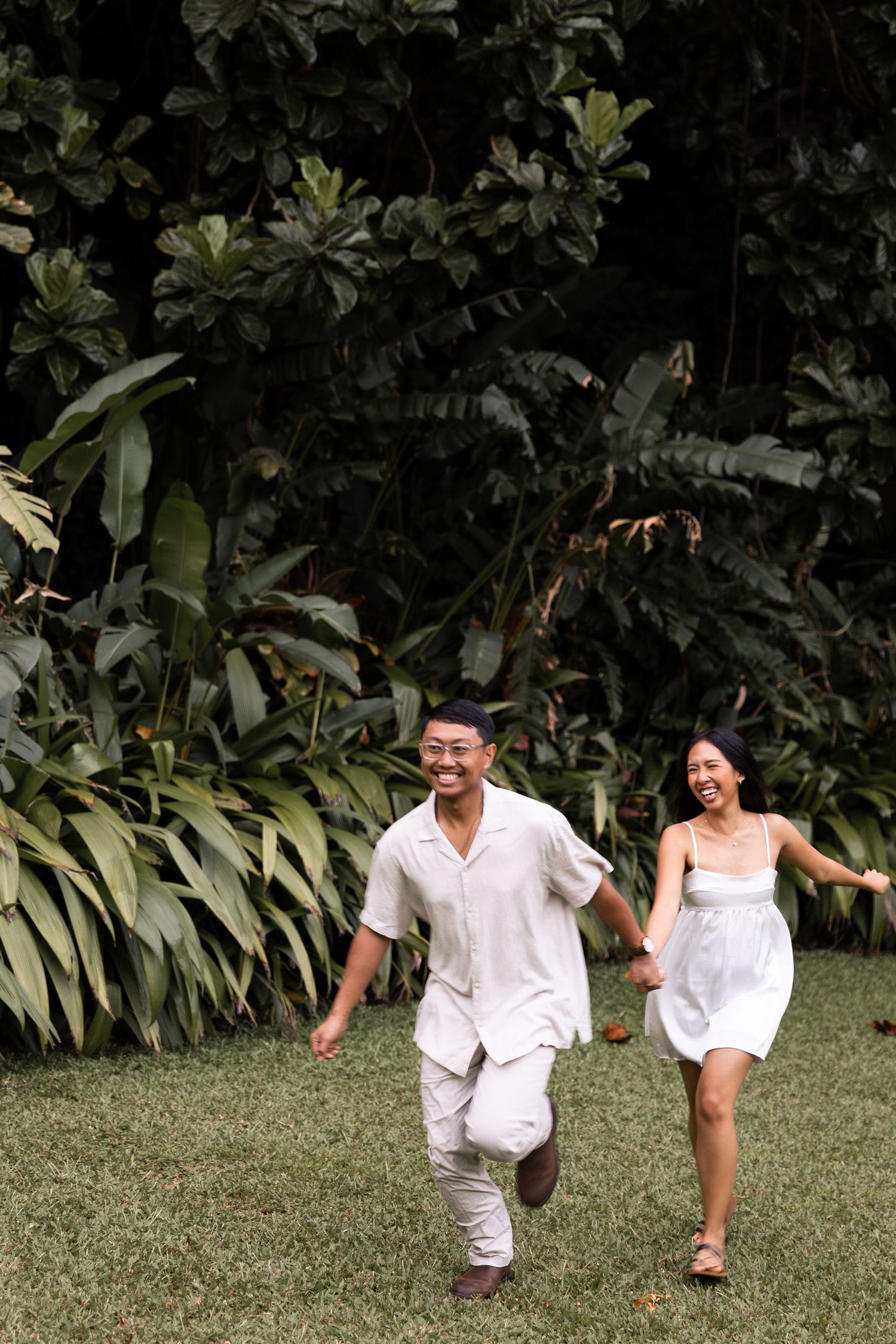 Couple in white attire running joyfully through a garden with tropical foliage.