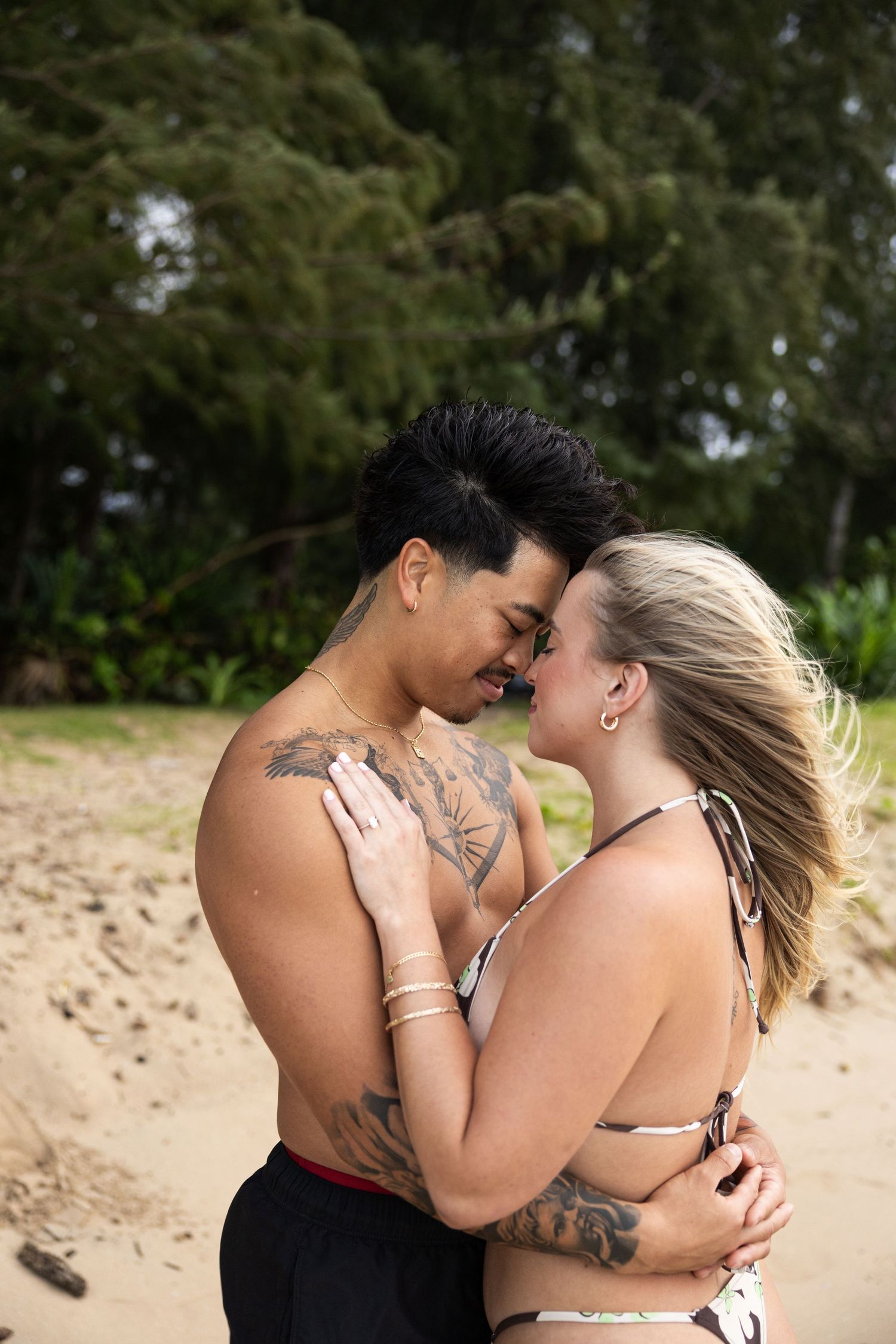 A couple shares an intimate embrace on a tropical beach with mountains and trees in the background.