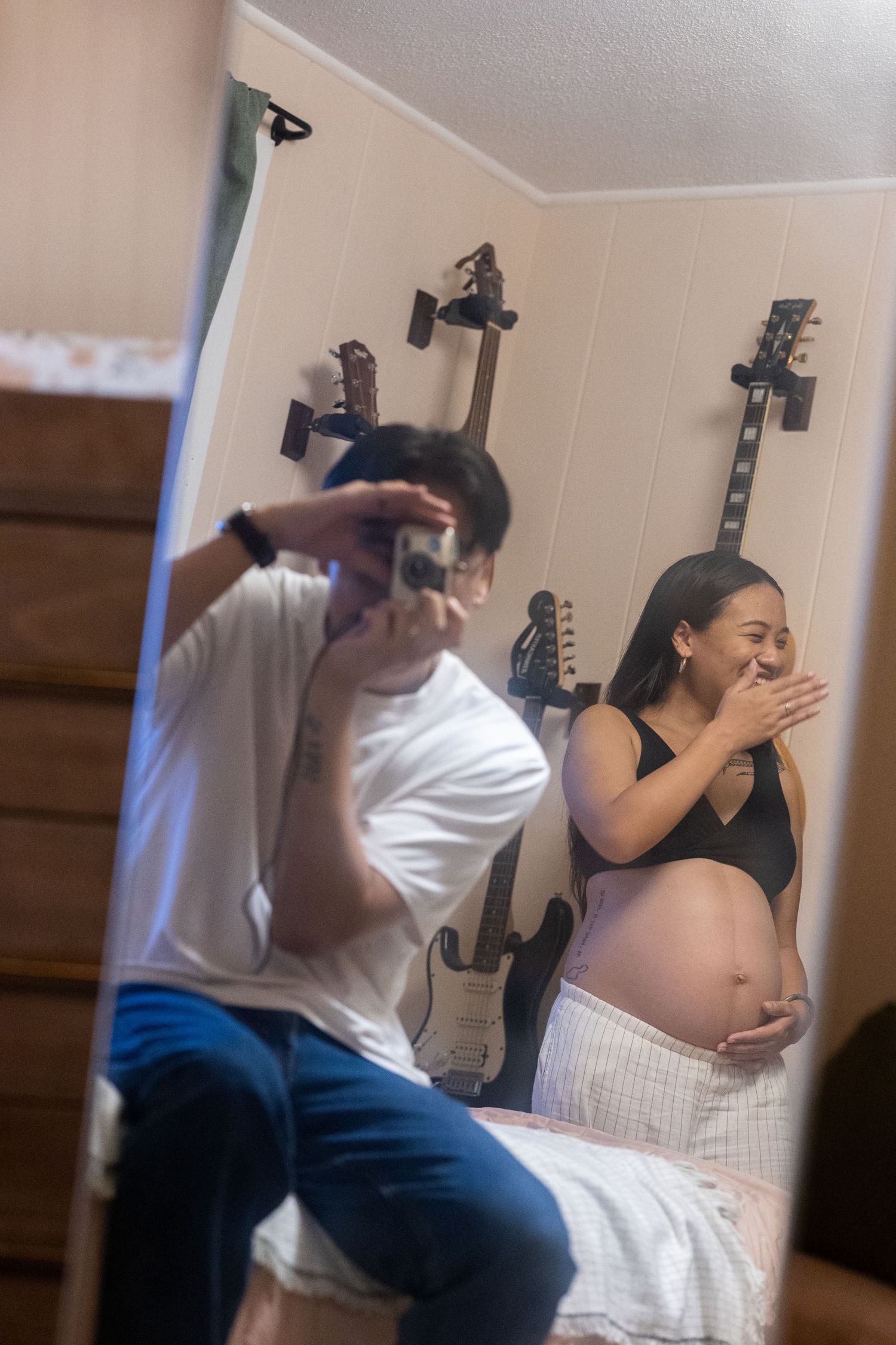 A maternity photoshoot moment with two people reacting playfully in a bedroom with guitars on wall.