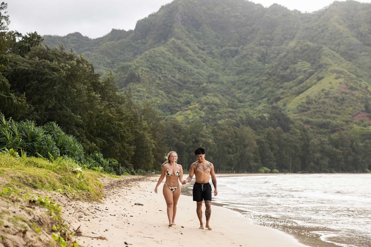 A couple walks together along a scenic Hawaiian beach with dramatic mountain backdrop.