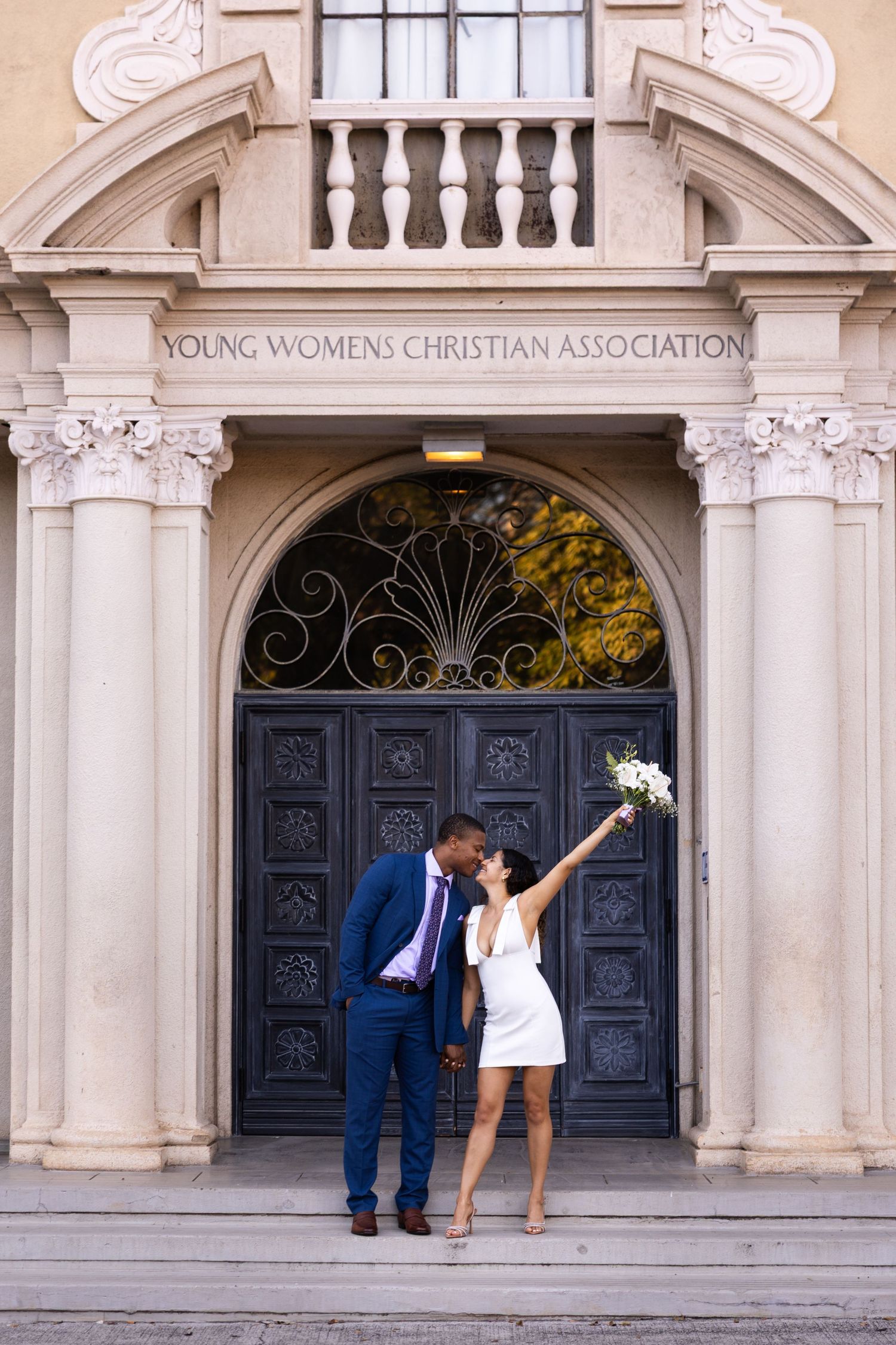 A couple poses in front of an ornate YWCA building entrance with decorative columns and wrought iron doors.