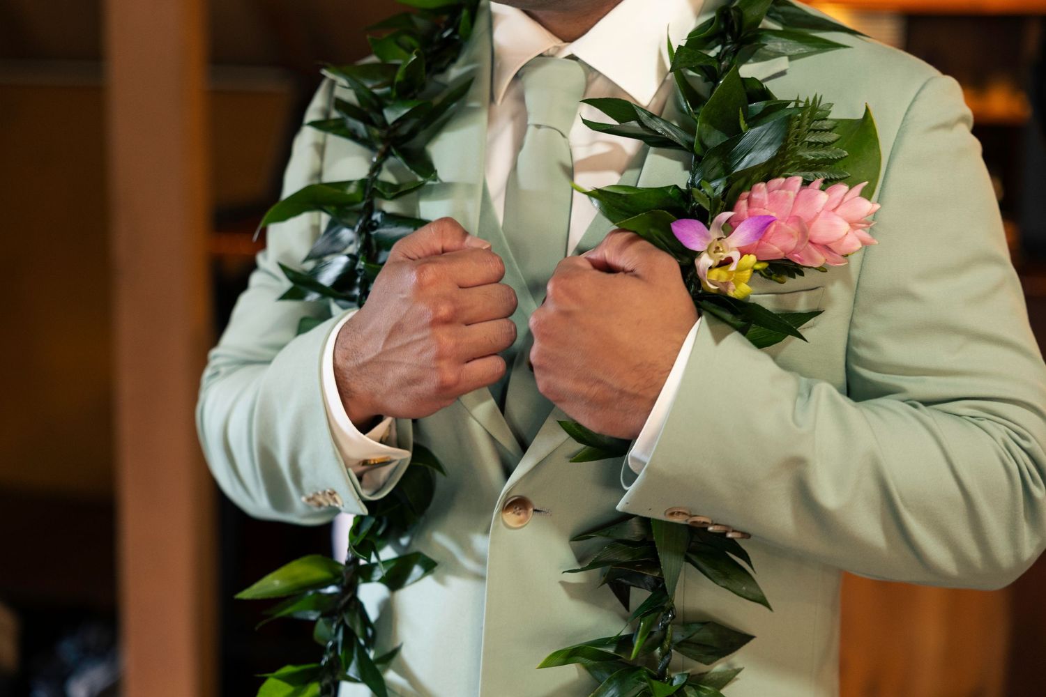 Person in light suit adjusts boutonniere flower arrangement against wooden backdrop.