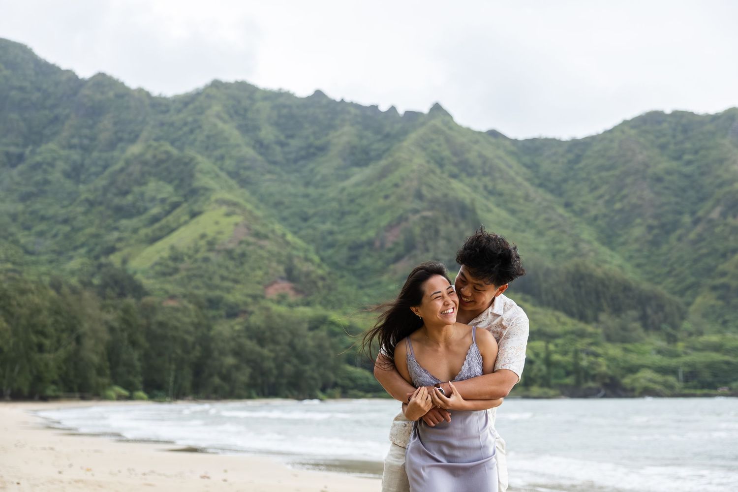 Couple sharing a tender moment while walking on a tropical beach with dramatic mountain landscape.