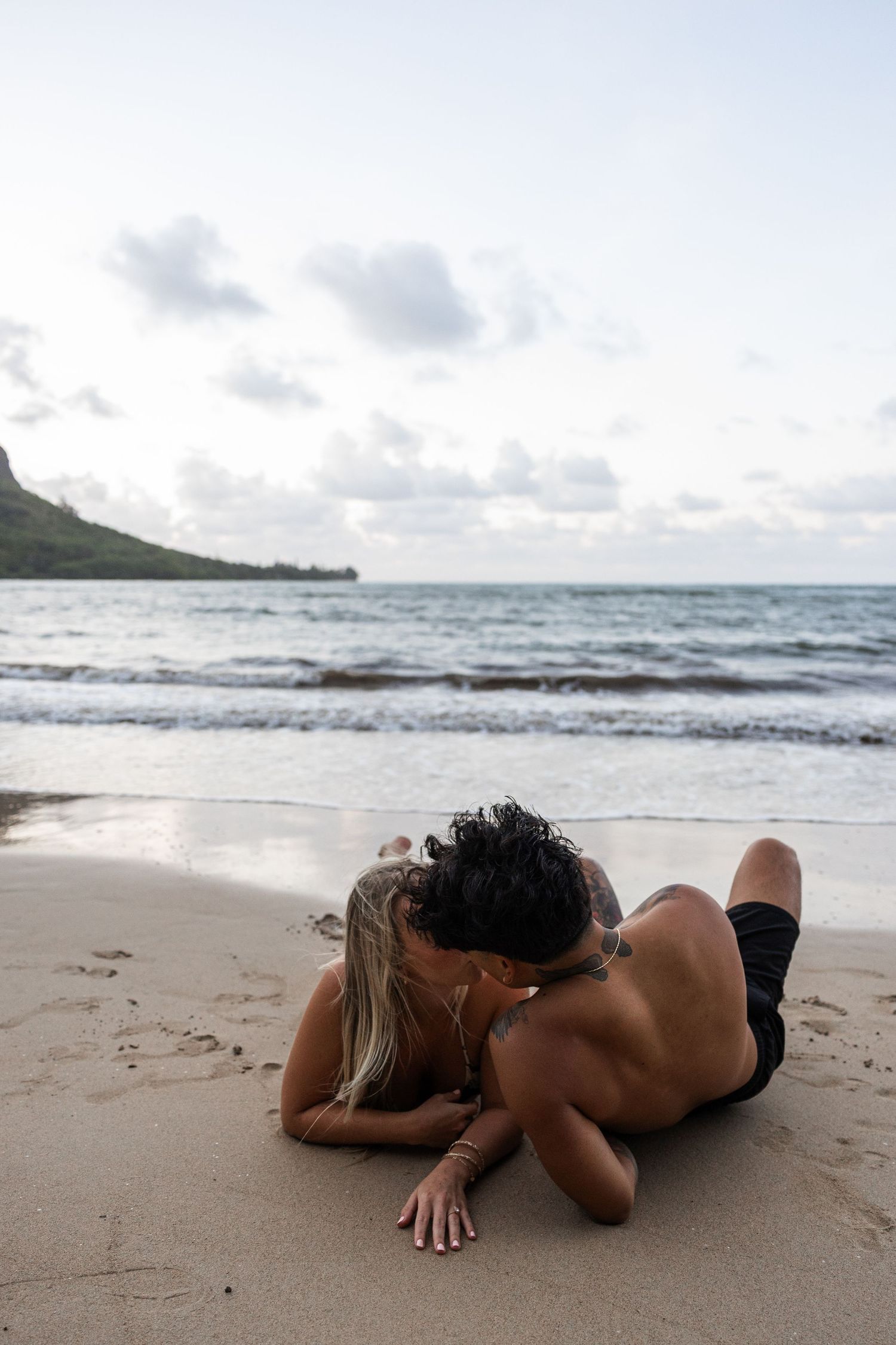 Person lying on sandy beach with waves and mountains in background during cloudy day at shoreline.
